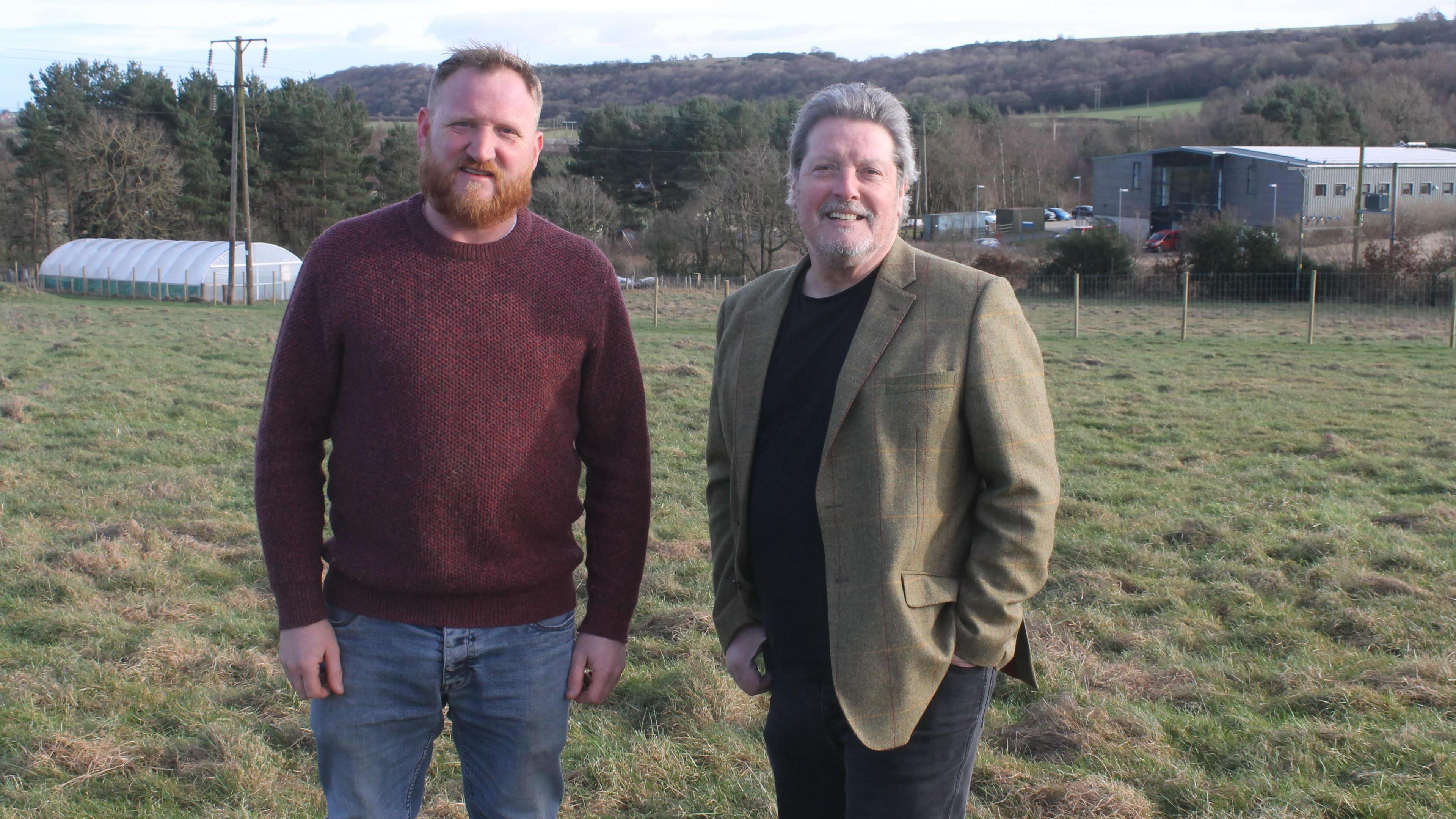 Dave Wilde, who has a ginger beard, is wearing a claret jumper and blue jeans. John Phillipson is wearing a green/brown blazer and a black round neck top with dark trousers. They are standing next to each other in field.