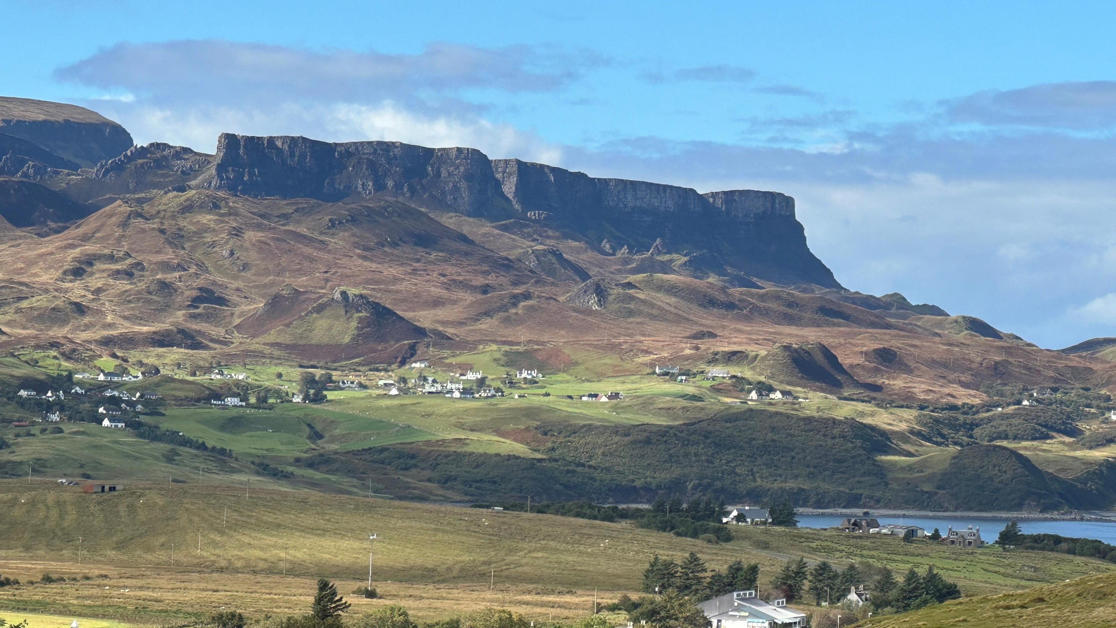 A picturesque countryside view with steep hills and houses dotted around and a small loch in the corner