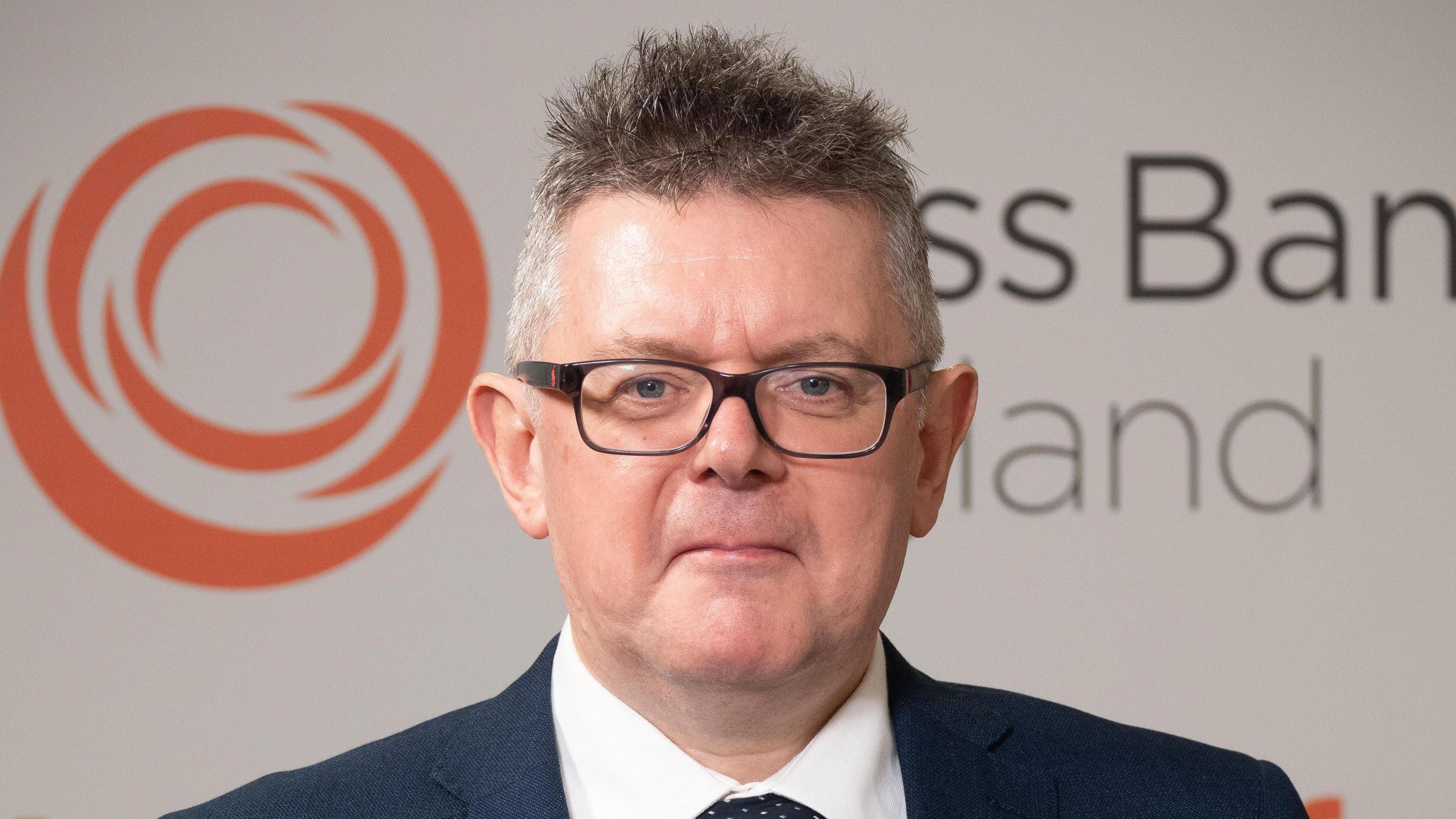 A man with short grey hair and glasses wearing a suit standing in front of an orange and white Brass Band England logo.