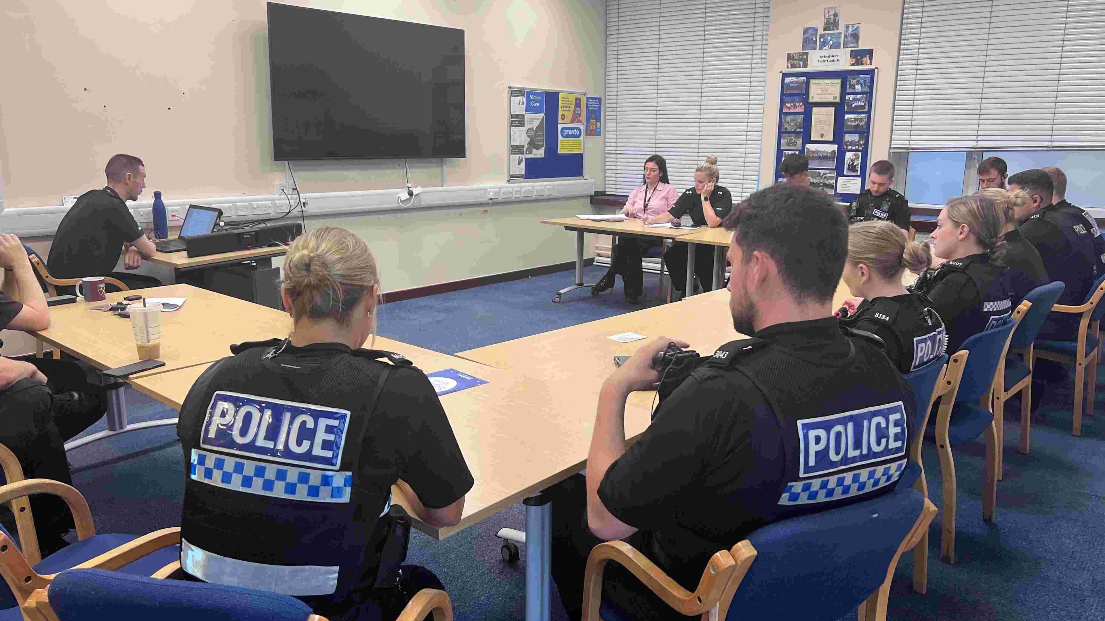 A group of police officers, in a room. They have "police" written on their back, and there is a blue carpet, a number of tables and a large television screen. 