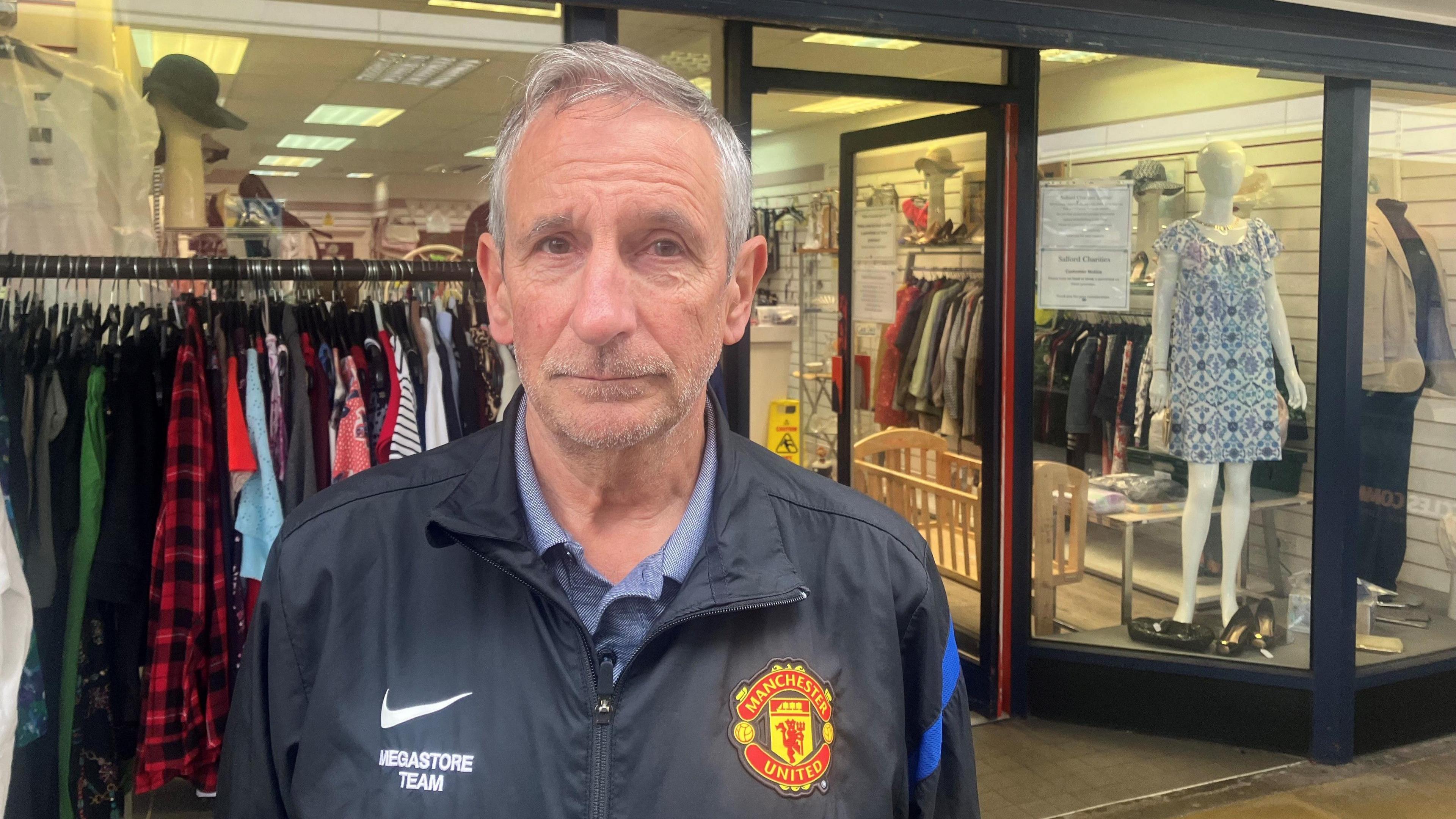 Barry Fallows stands outside of his charity shop. He is not smiling and wears a black jacket with a Manchester United logo. Racks of clothes and a shop window with mannequins with clothes on are in the background