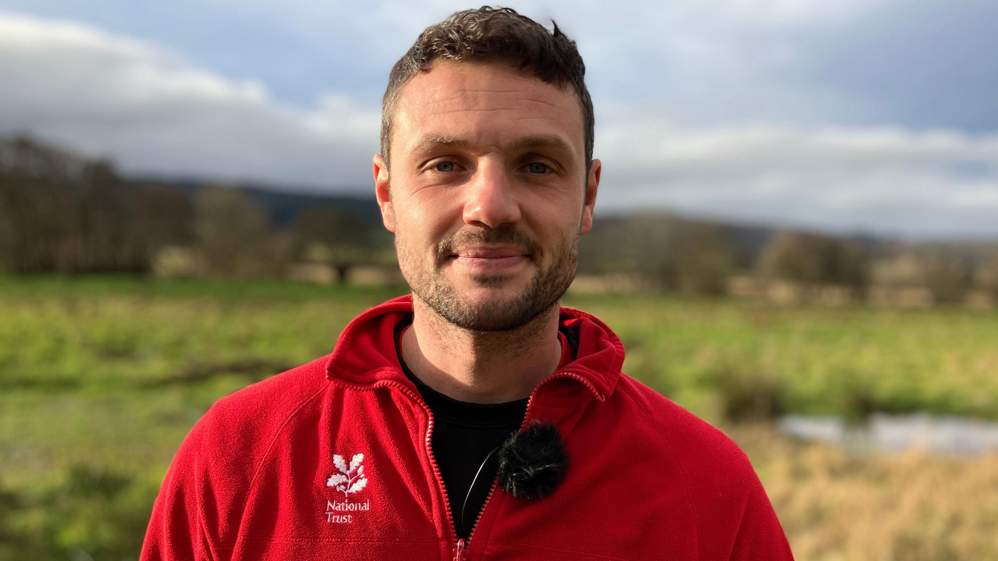 Jack Siviter. He is pictured outdoors in Holnicote Estate. The landscape is out of focus but green fields and trees can be seen behind him. Jack has short dark hair and facial hair. He is wearing a red fleece with a white National Trust logo on the chest. He is looking directly at the camera and smiling 