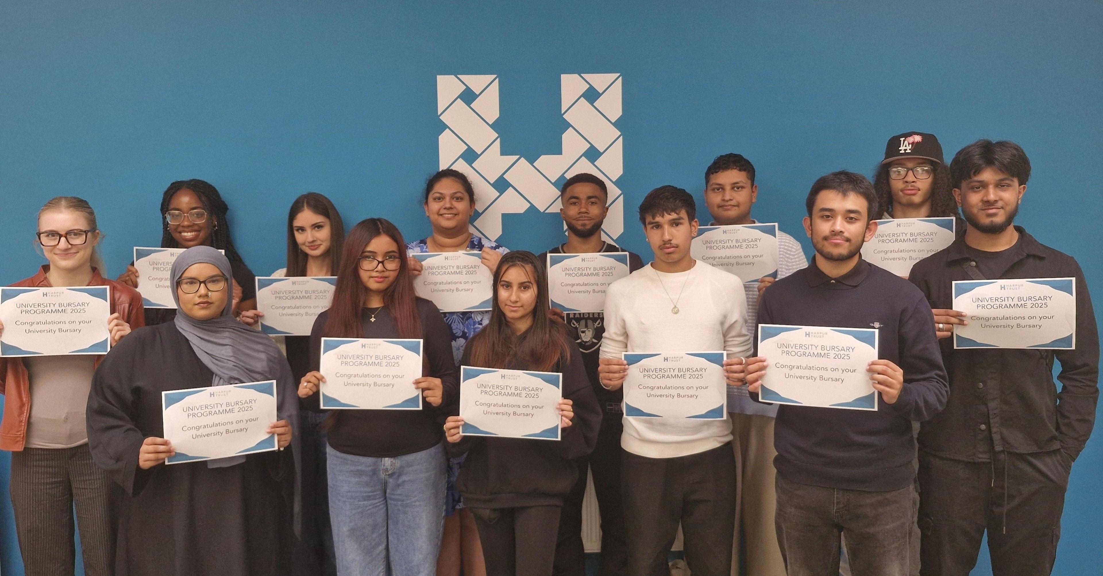 Thirteen students, standing by a blue wall, holding a certificate, all looking at the camera. 