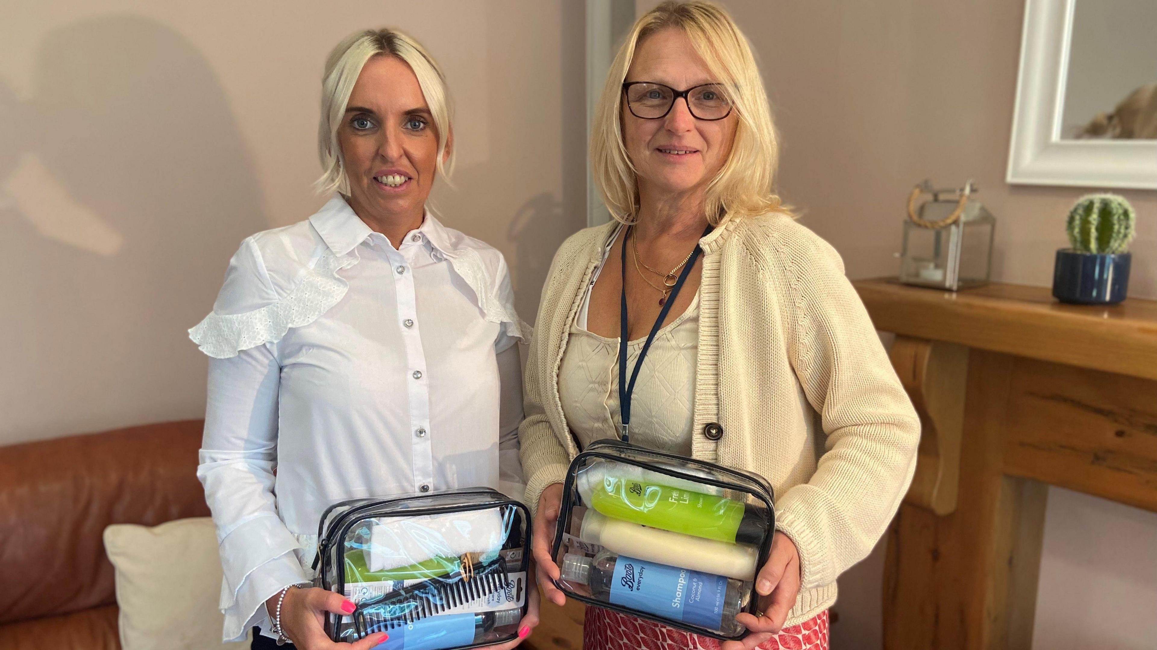 Katie Donnelly and Julie Boner standing in a room. Both are holding the wash bags which are clear and are filled with personal care items