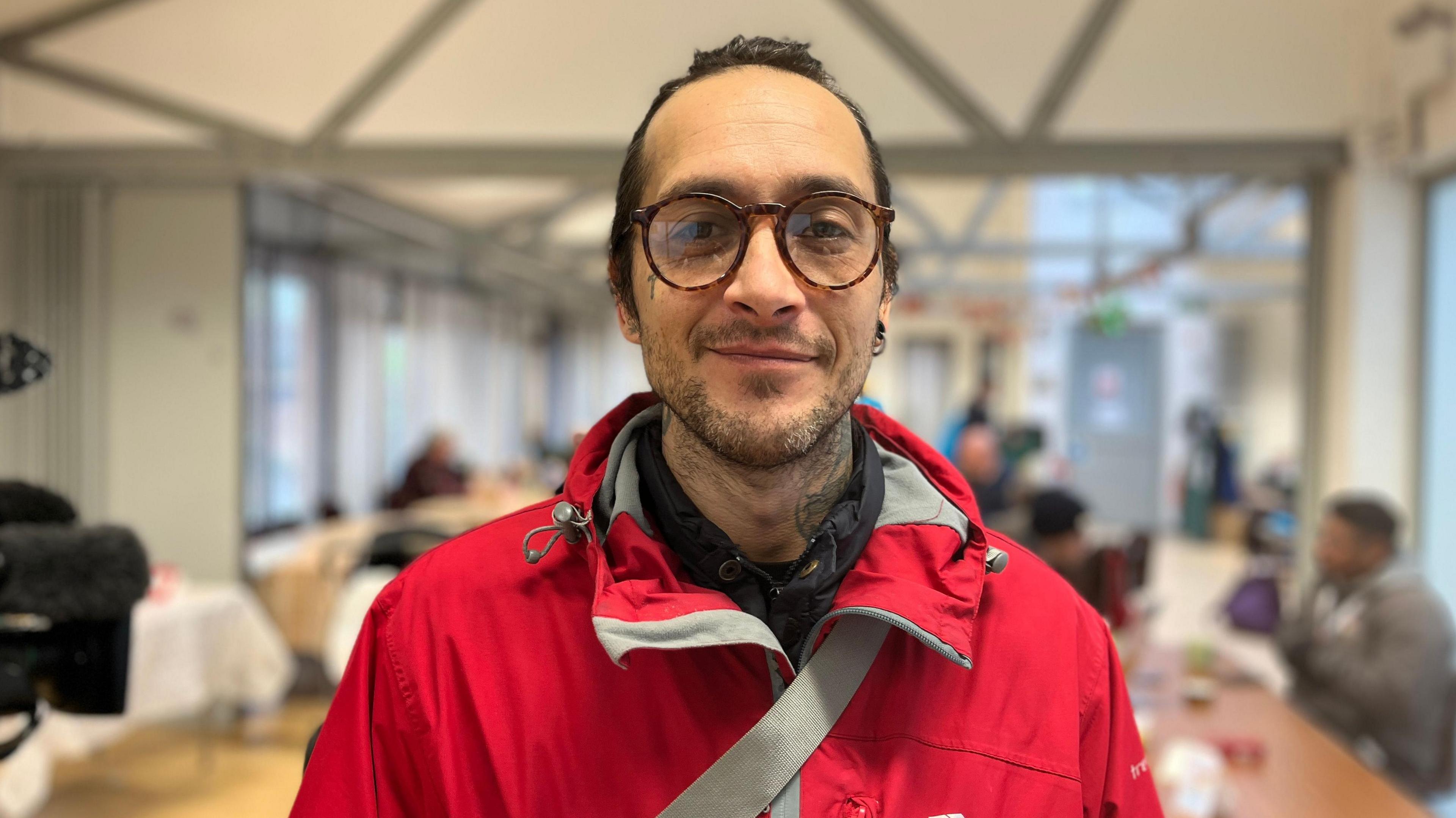 Tony Hill is wearing brown rimmed glasses and is looking straight at the camera. he is wearing a red and grey jacket with a hood.