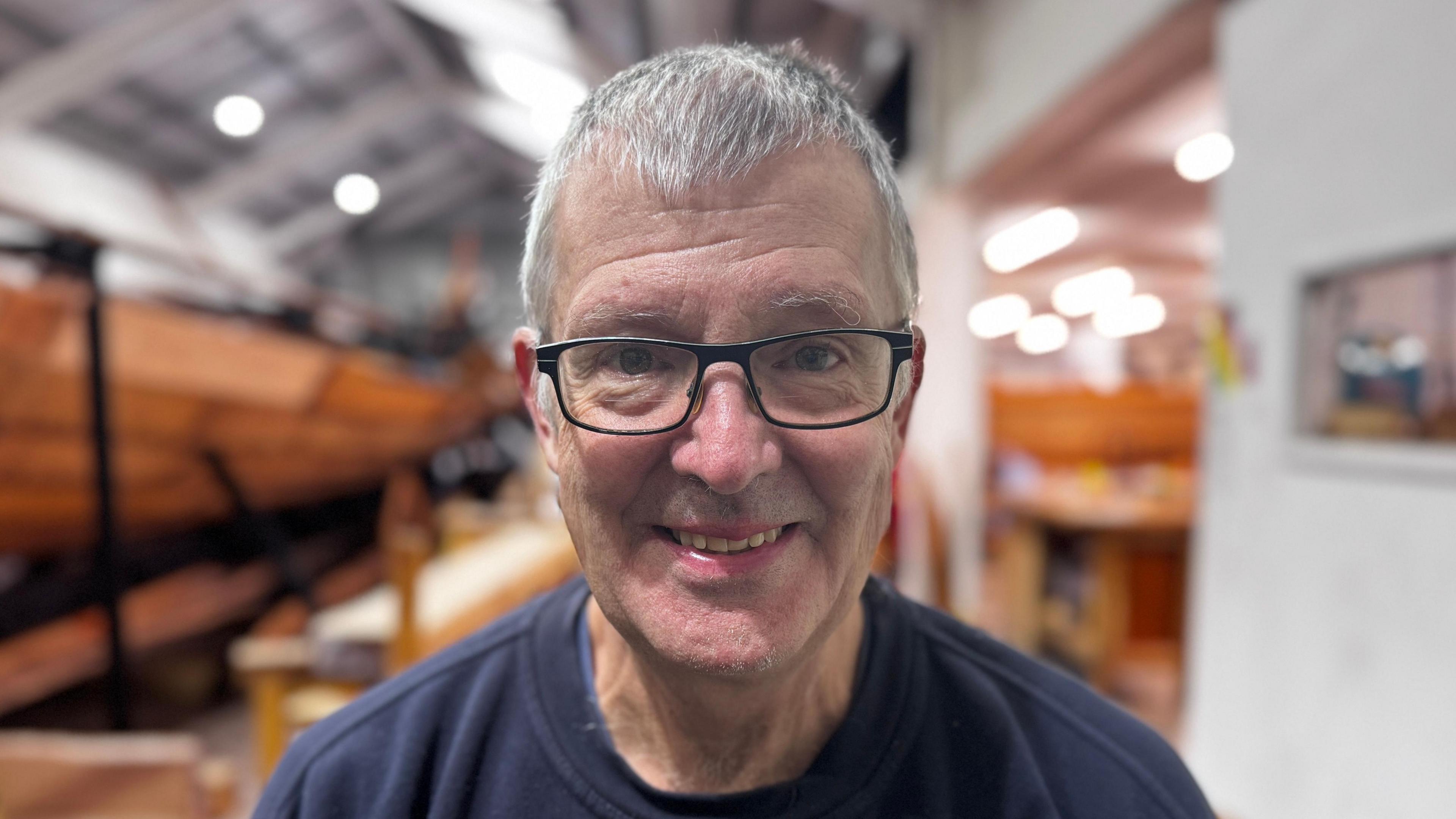 Dave Clemmer stands in front of the ship reconstruction and smiles at the camera. He has short grey hair and wears a navy jumper and glasses. 