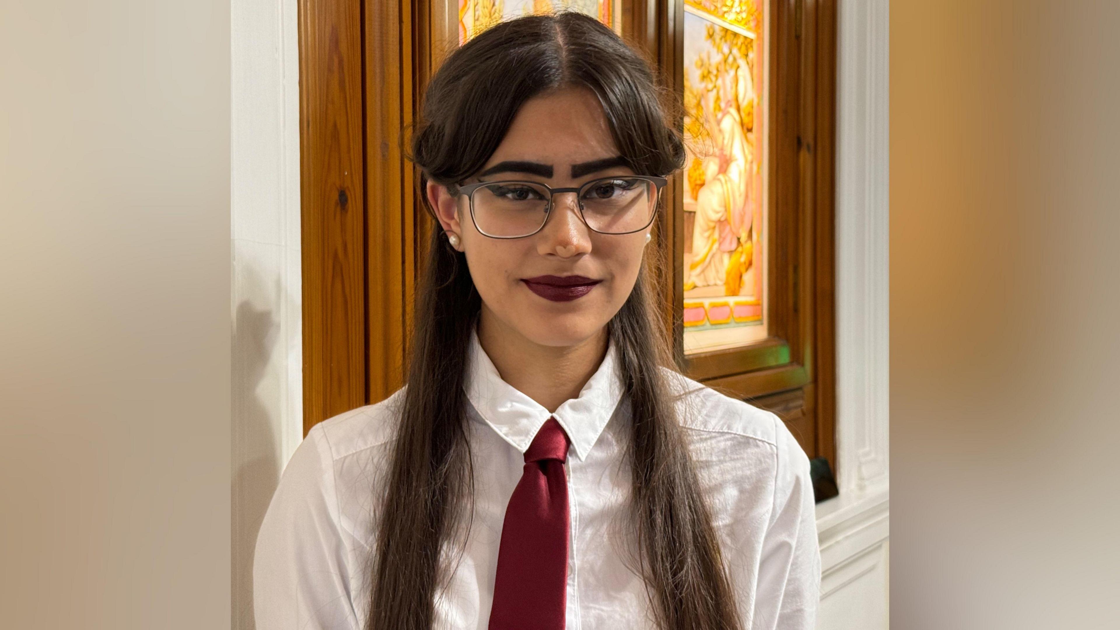 Malia Dayas, who has long brown hair and glasses, is wearing dark lipstick, a white blouse and ruby-coloured tie. She stands in front of a stained glass window.