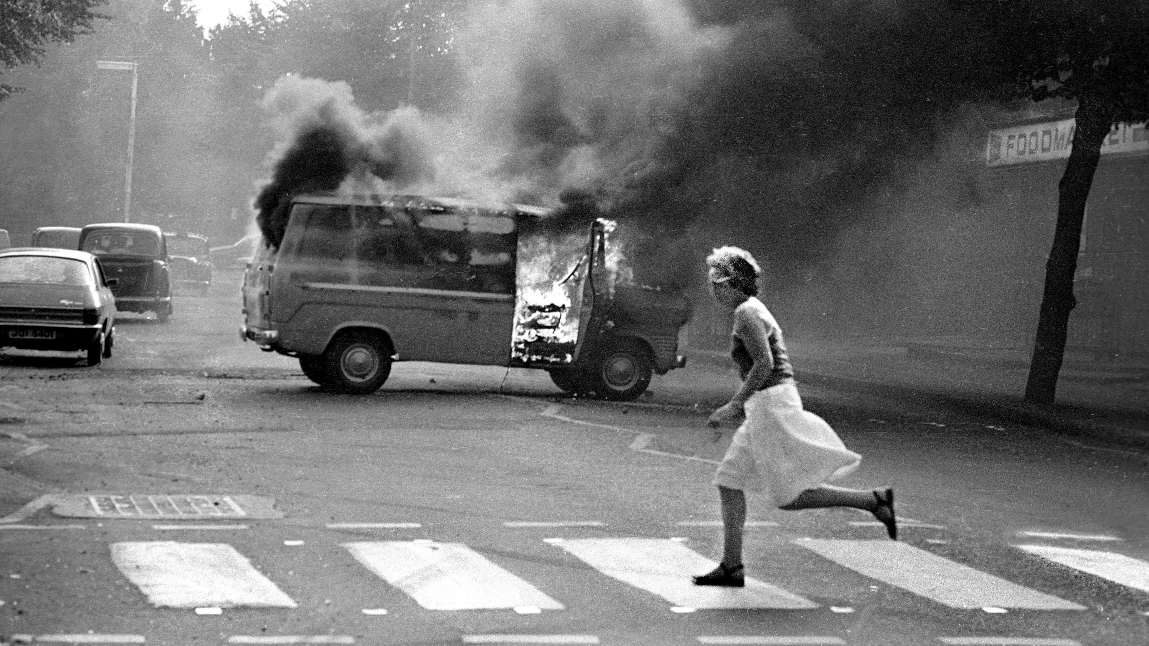 A black and white image of a vehicle burning out as a woman runs across a pedestrian crossing. There are cars driving past the burning vehicle, which is filling the air with smoke.