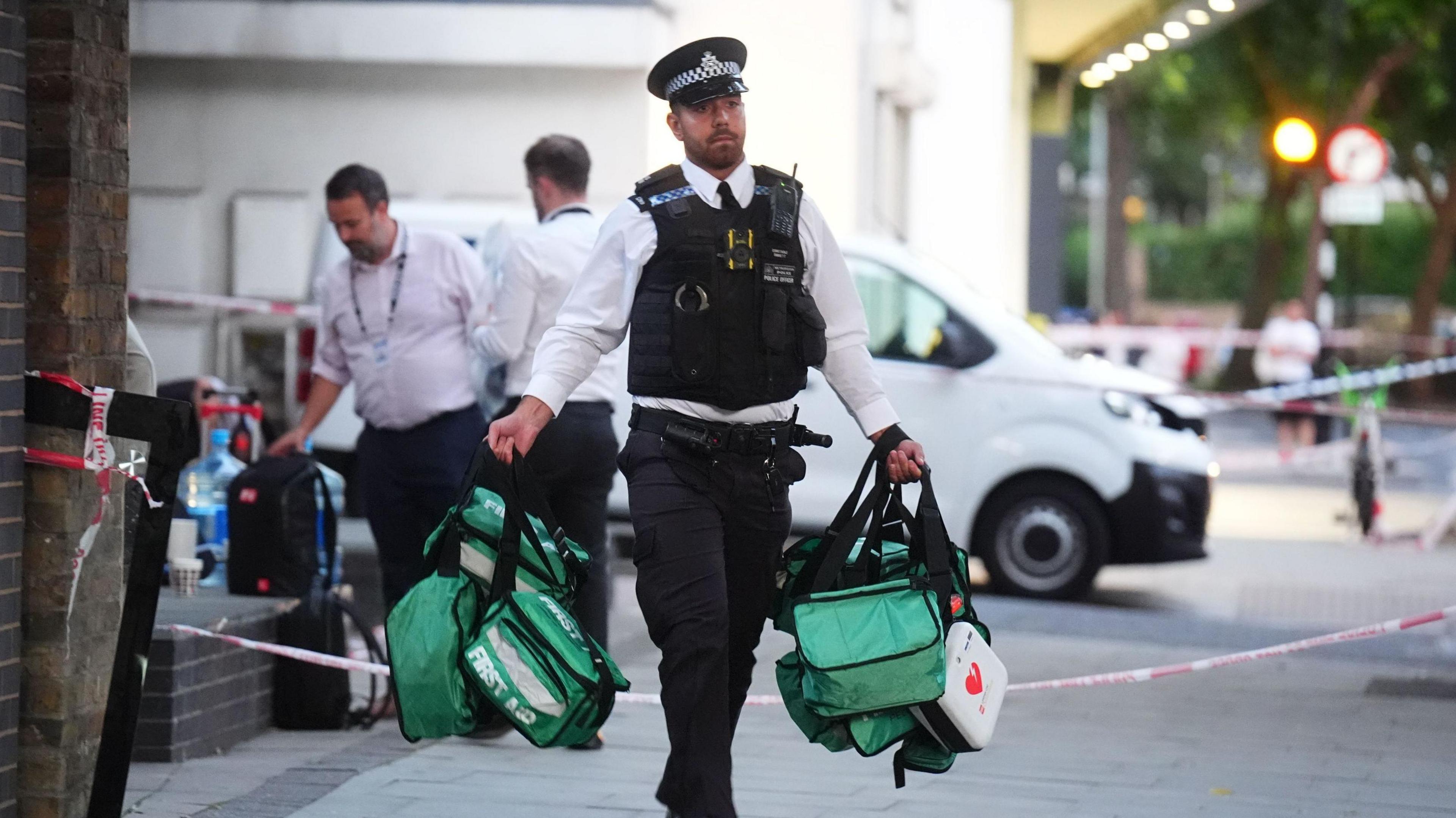 A police officer carries green first aid bags away from the scene of the stabbings. Police tape blocking the road is visible behind him.