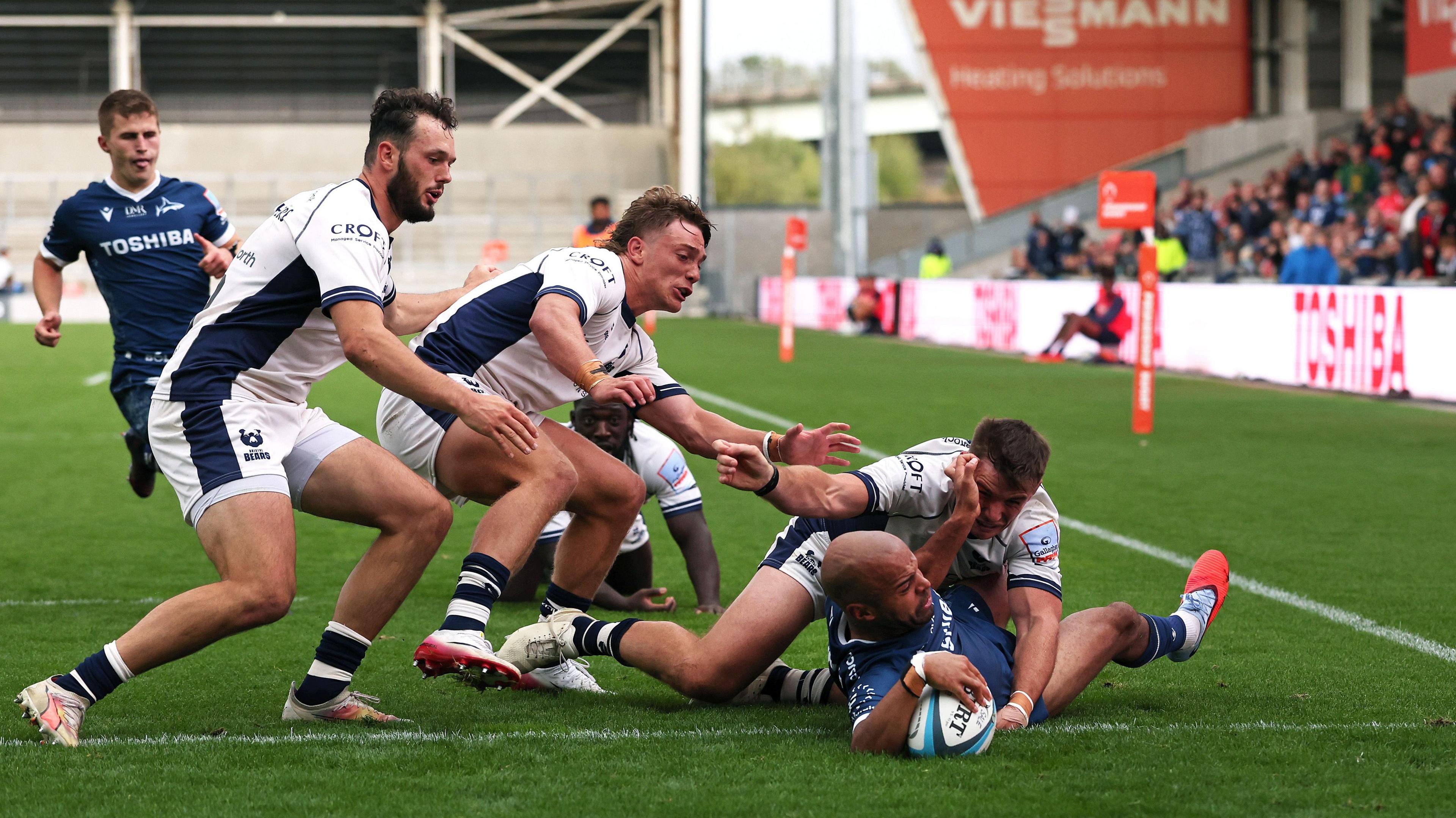 Sale's Tom O'Flaherty, wearing navy blue, puts the ball down over the try-line despite the attention of three Bristol players in white. A team-mate watches him score in the background.
