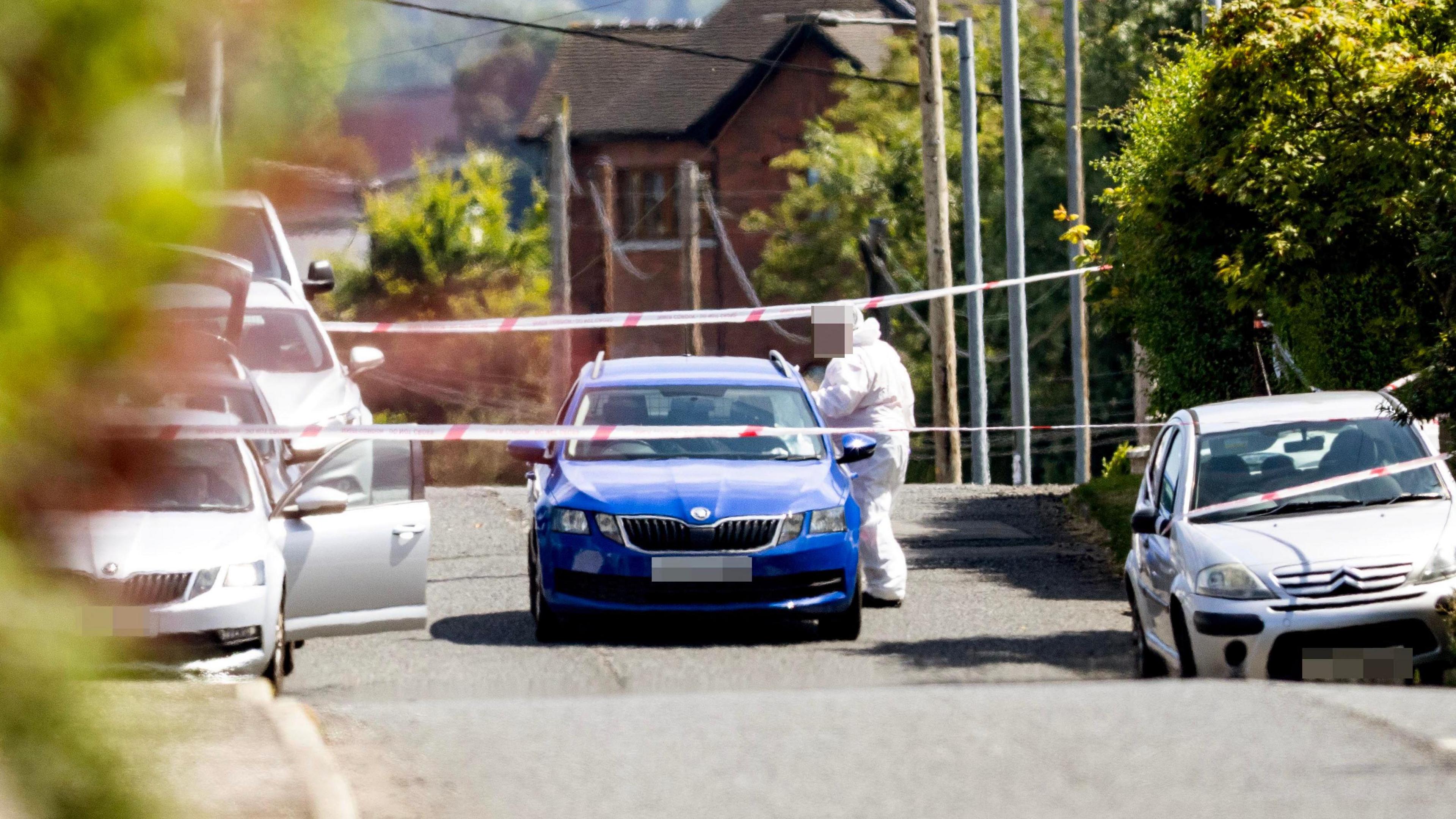 A forensic officer stood beside a blue car, the road is taped off with multiple white cars parked either side