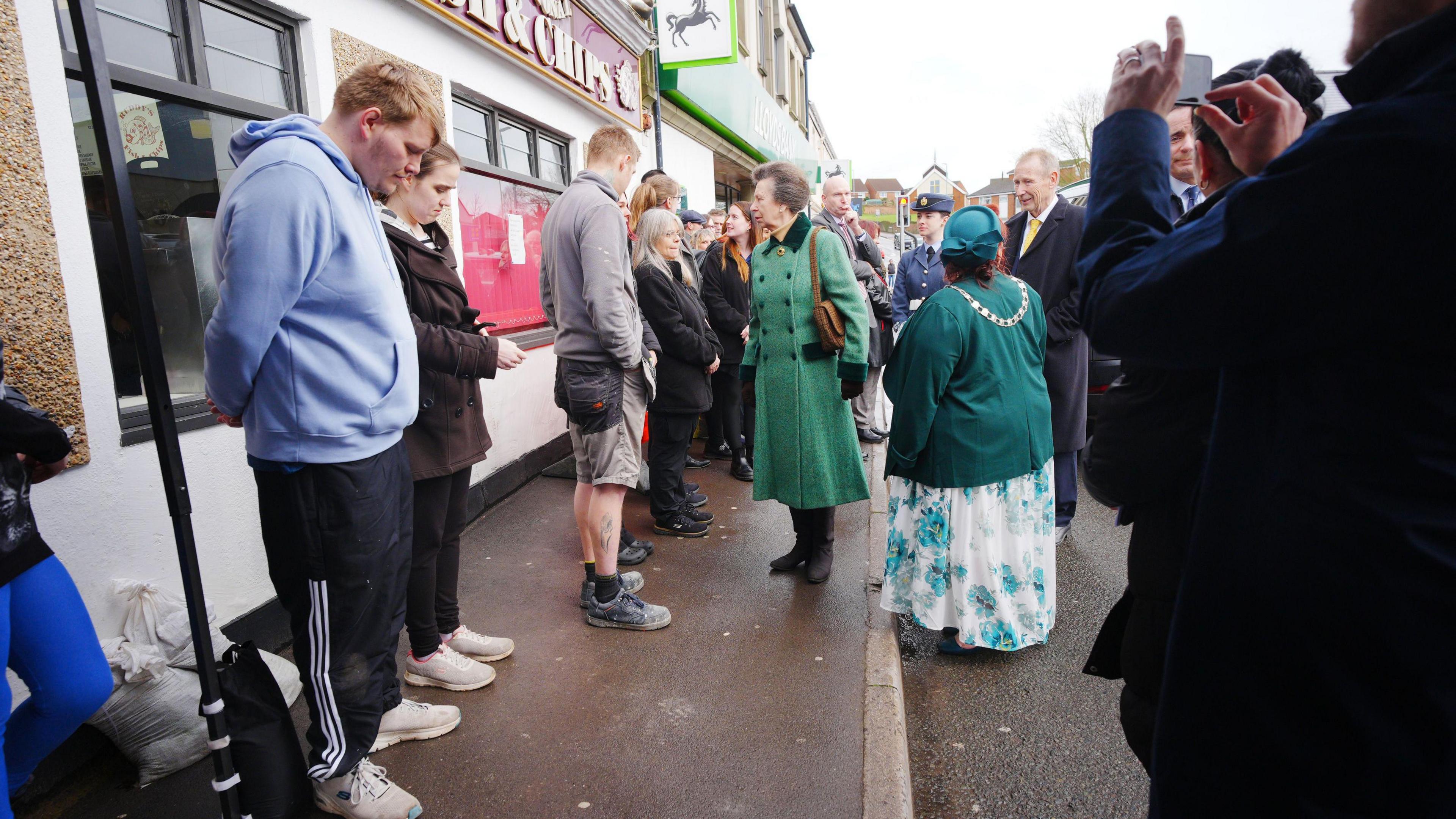 Princess Anne wearing a long green coat stands outside of a fish and chip shop. There is a row of people in front of her, and one person taking a picture.