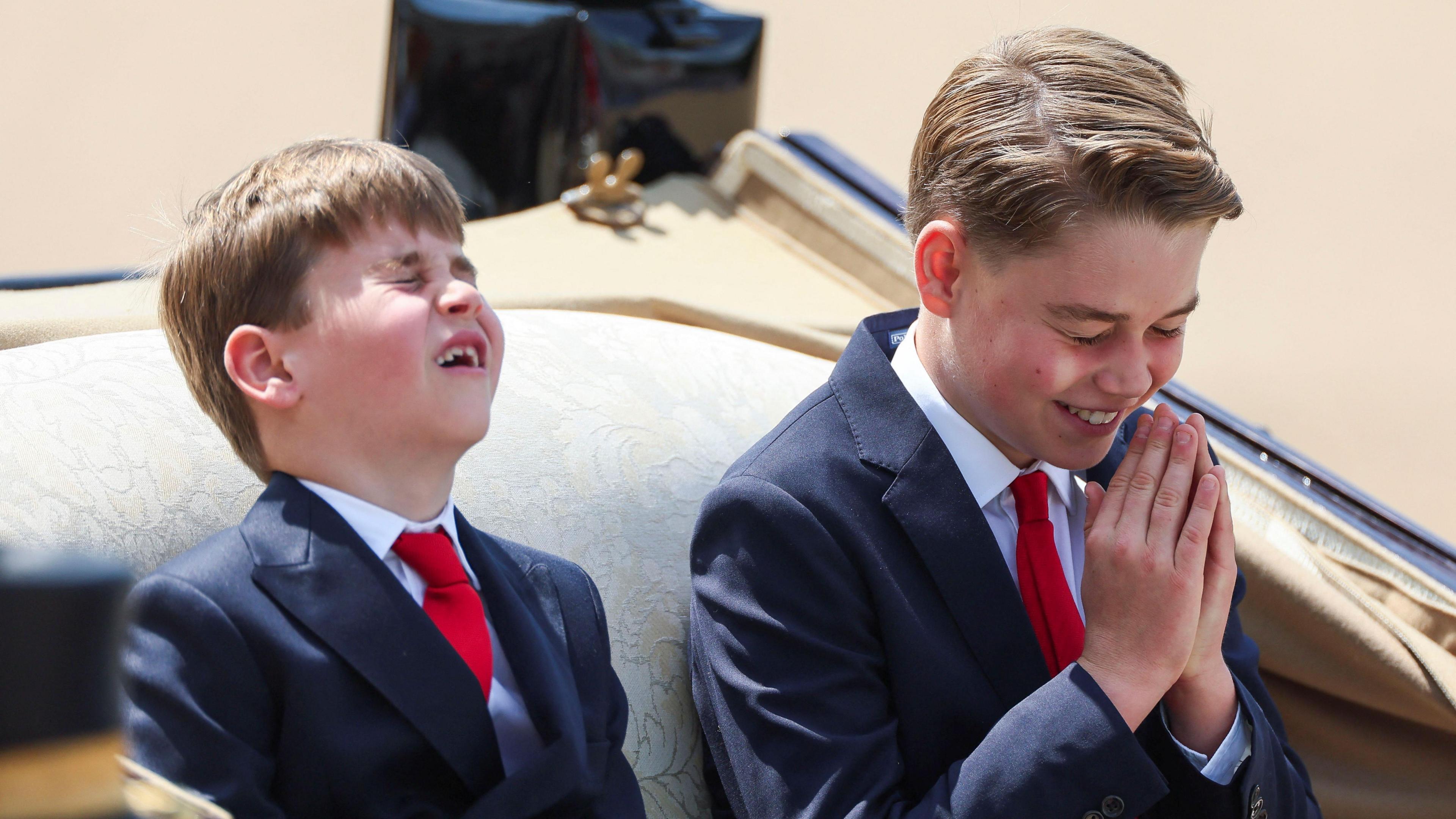 Prince Louis and Prince George ride in a carriage. George is laughing with his hands raised over his mouth
