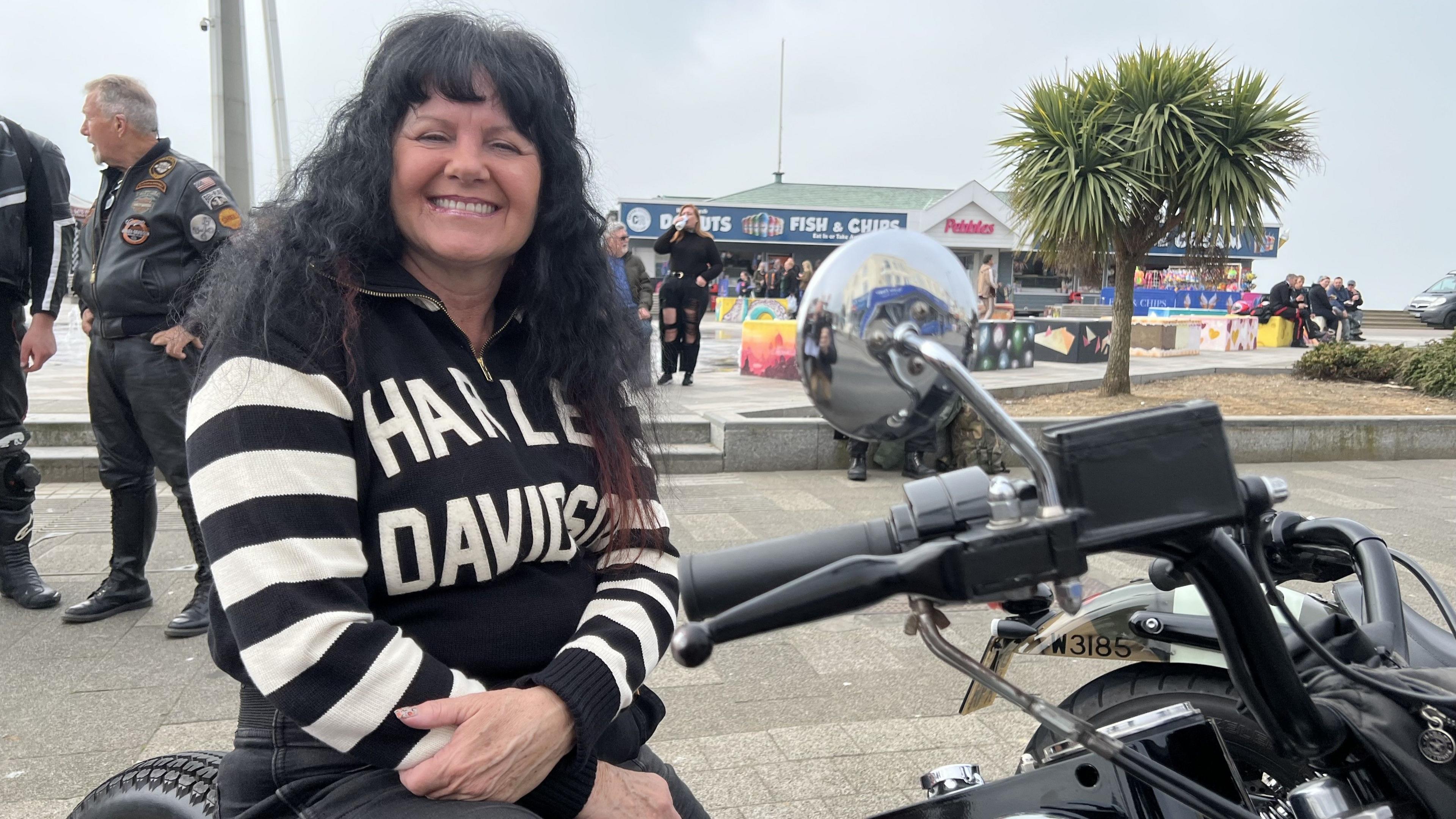 Karen Gelsthorpe smiles at the camera as she sits on her Harley-Davidson motorbike. She has shoulder length dark hair and wears a black jumper with the words Harley Davidson written on the front, the arms are white and black striped.