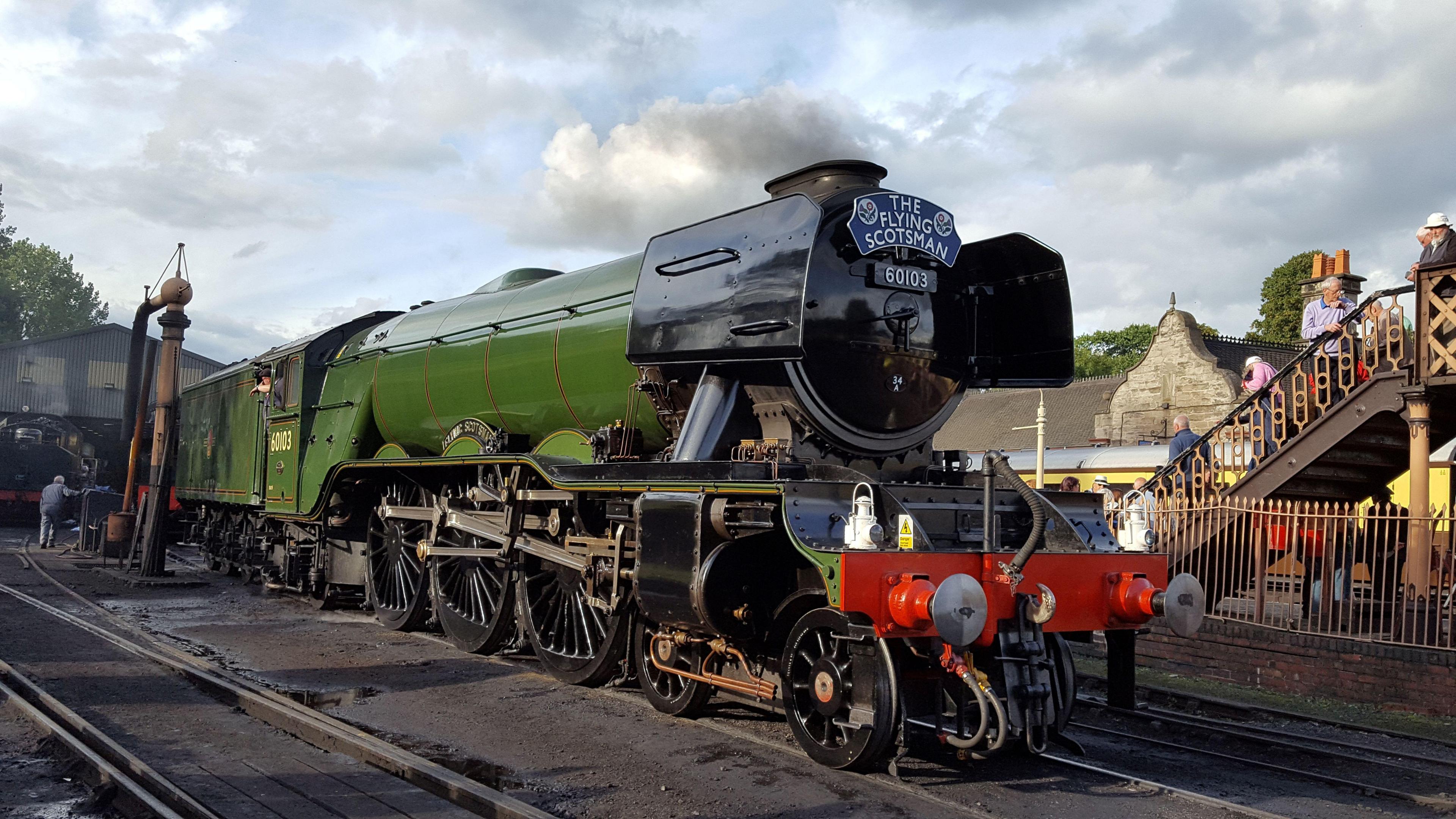 The front of the iconic Flying Scotsman locomotive stopped at a train station. It is green in colour with the very front face black with a blue sign at the top that says "The Flying Scotsman" and the number 60103 underneath. 