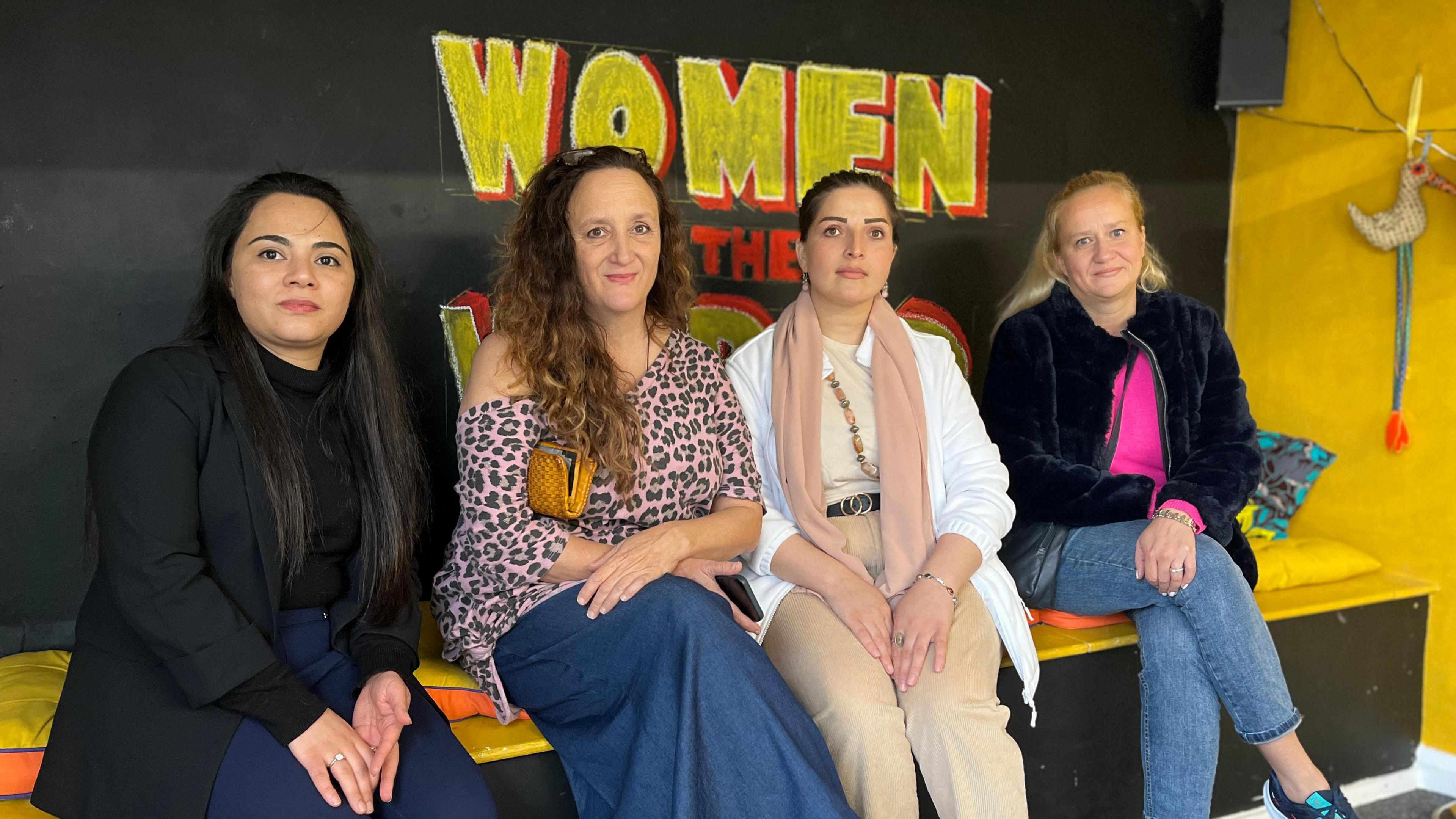 Saghar, Gillian, Sadaf and Olga sit on a bench in front of a chalk sign which reads Women of the World