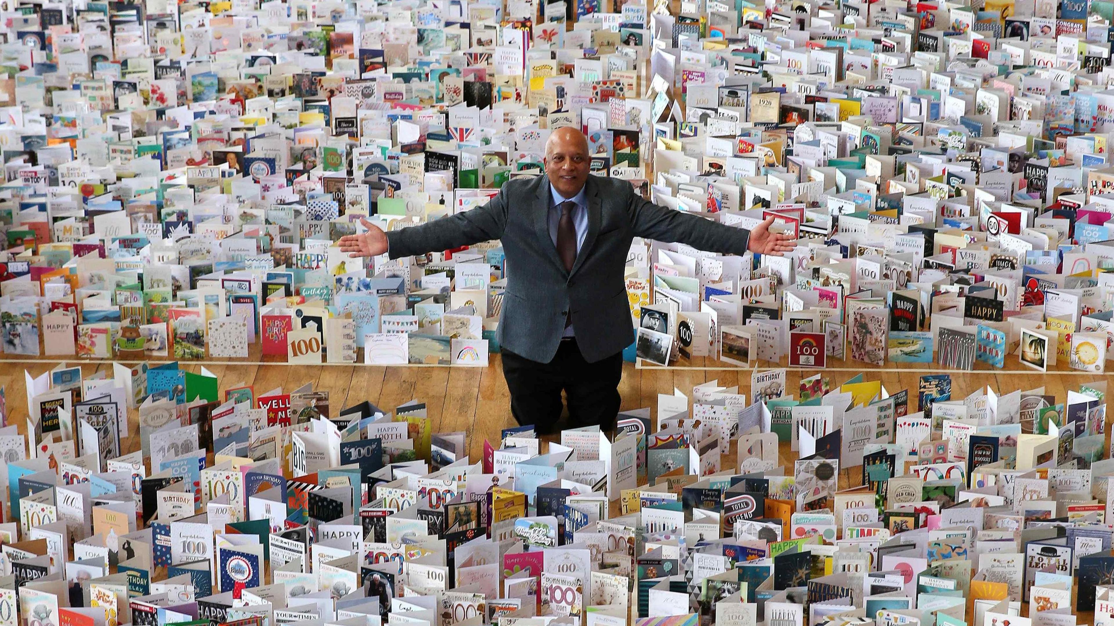 A man with his arms out standing amongst thousands of 100th birthday cards which are laid out on the floor