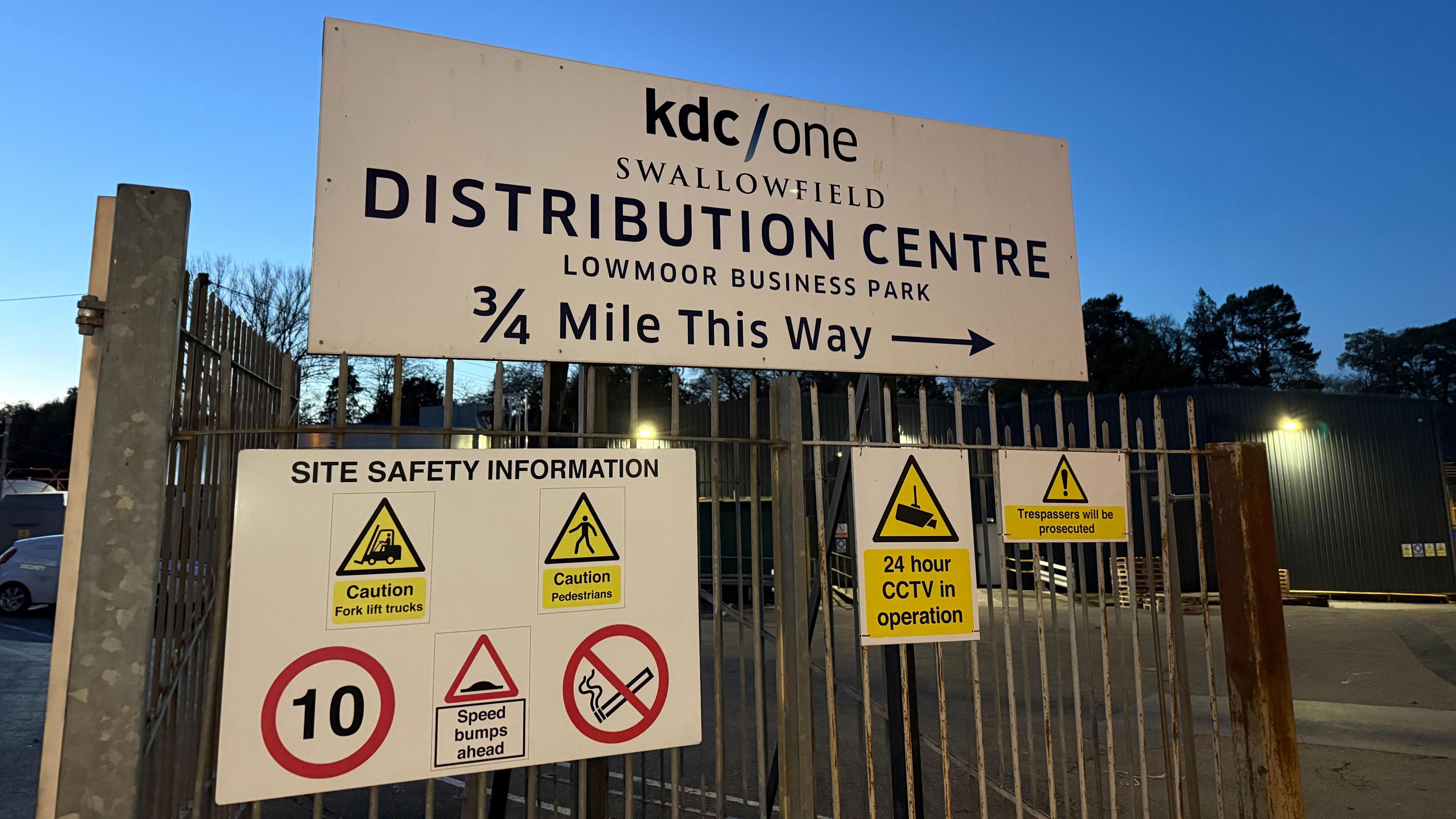 An entryway into the factory. There is a large sign on the gate which says 'KDC/One, swallowfield, distribution centre, lowmoor business park, 3/4 mile this way'. It is getting dark. 