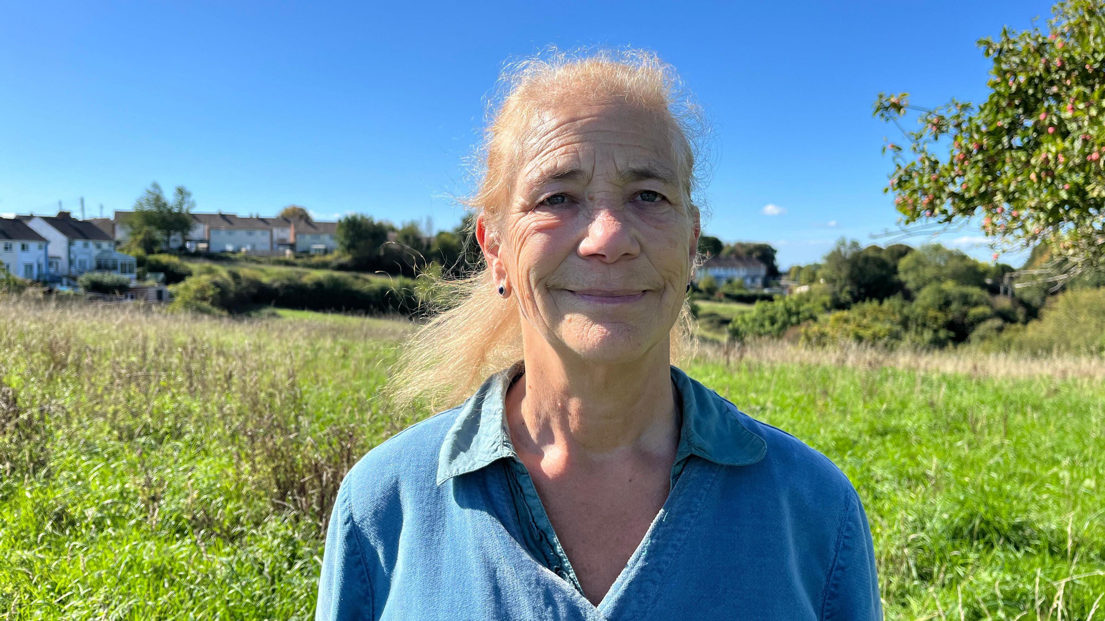 A woman in a blue top pictured from the chest up. She's looking at the camera and has a smile on her face. Behind her is a grassy orchard, and beyond that, houses.