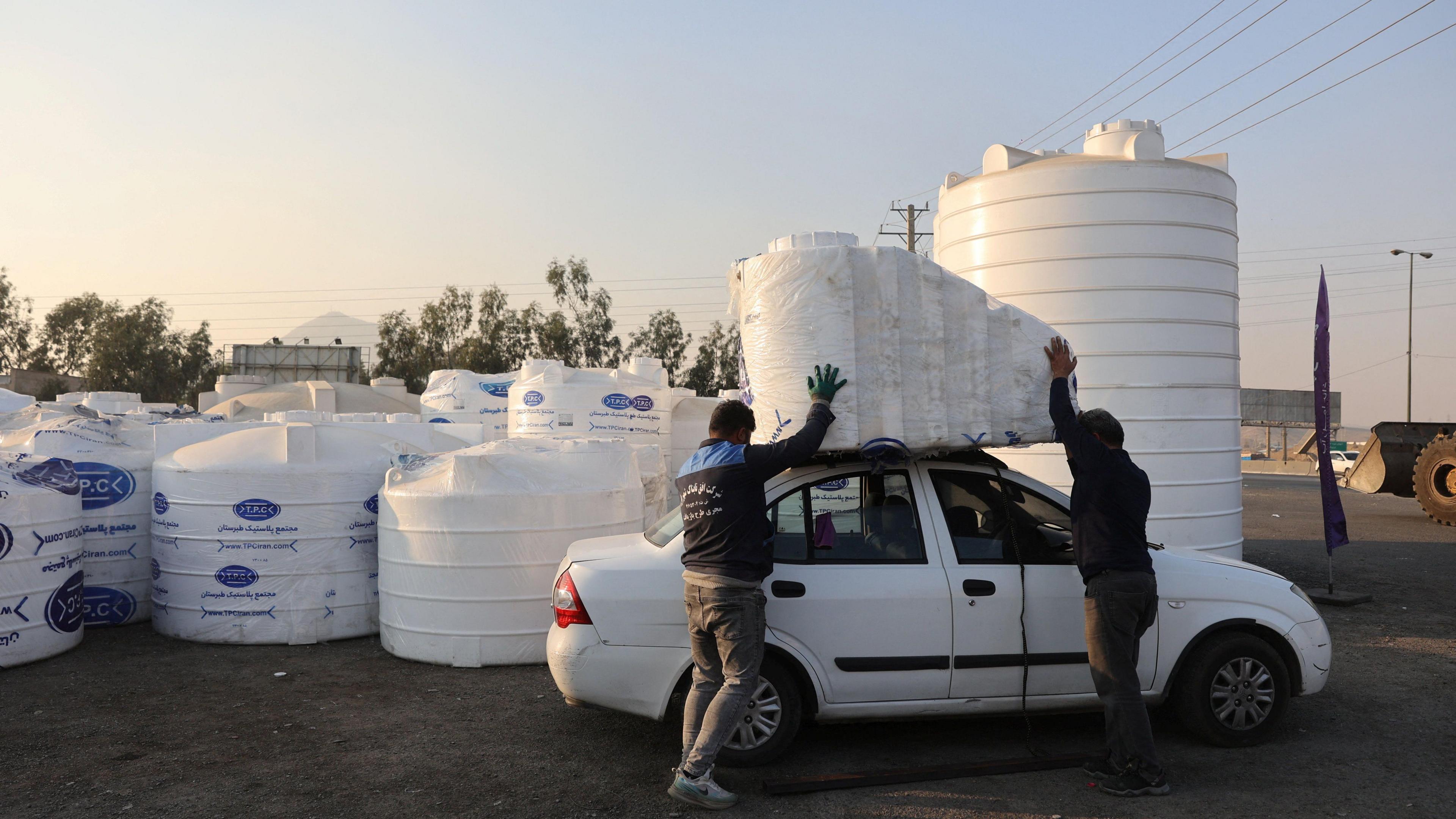 White plastic water storage tanks are seen with two people attaching one to the roof of a white car