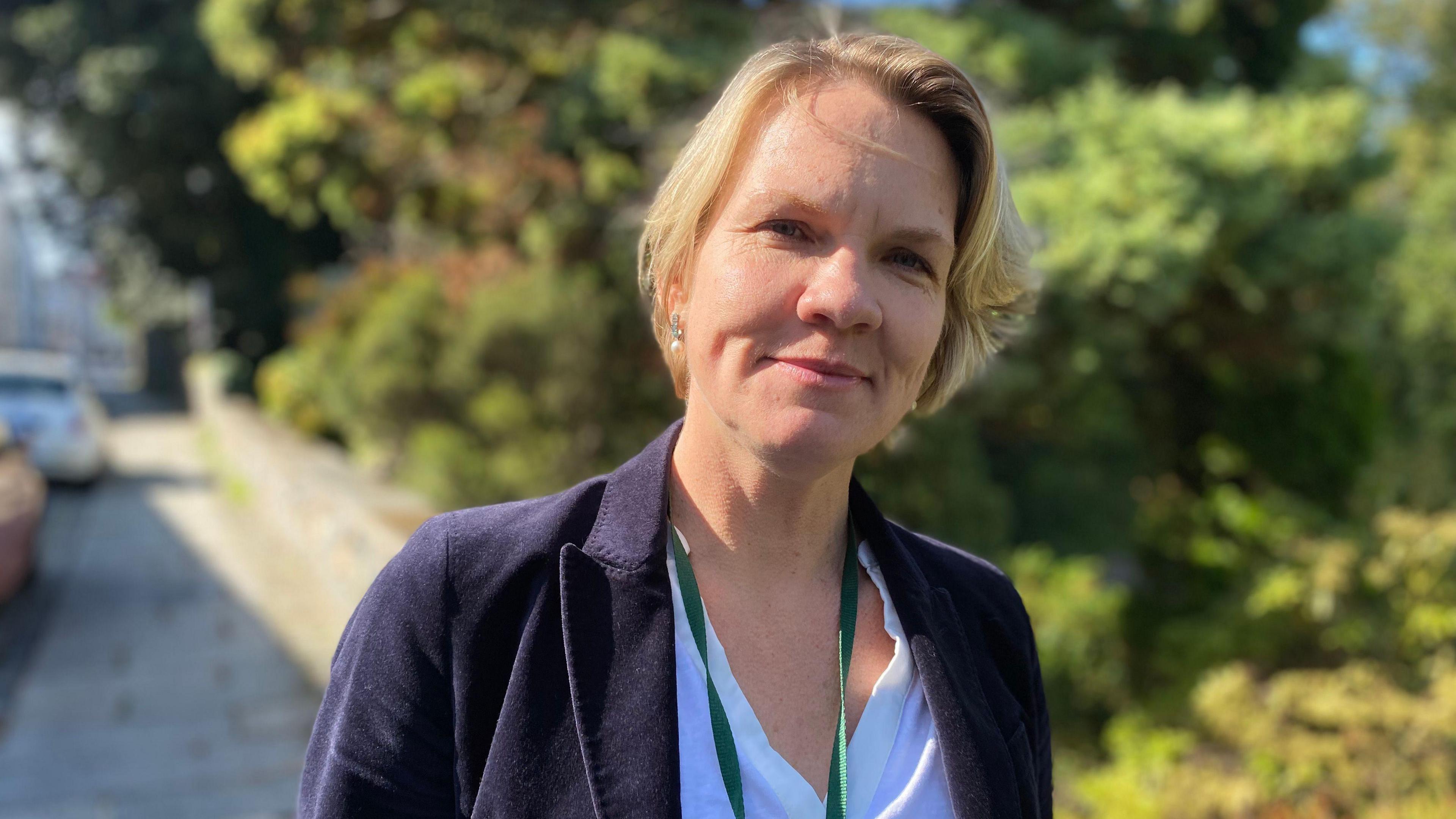 A gently-smiling woman with blonde, shoulder length hair in a white blouse and navy blue blazer, with a green lanyard on. She stands on a pavement with bushes and cars behind her on a sunny day.