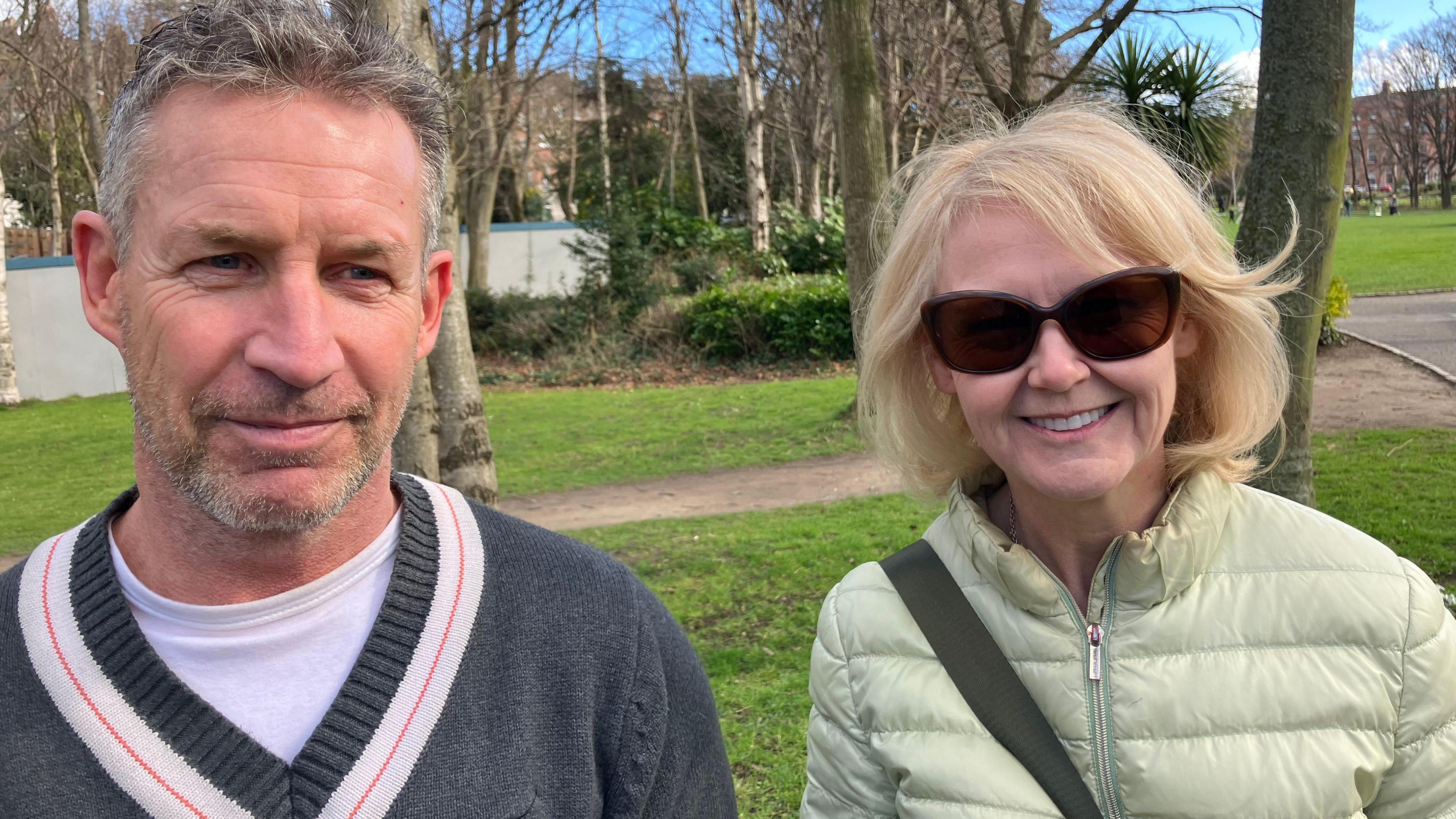 Les and Michelle in a park. There is grass and trees behind them. Les wears a grey jumper and white t-shirt. He has grey hair. Michelle wears a light green coat. She has blonde hair and is wearing sunglasses.