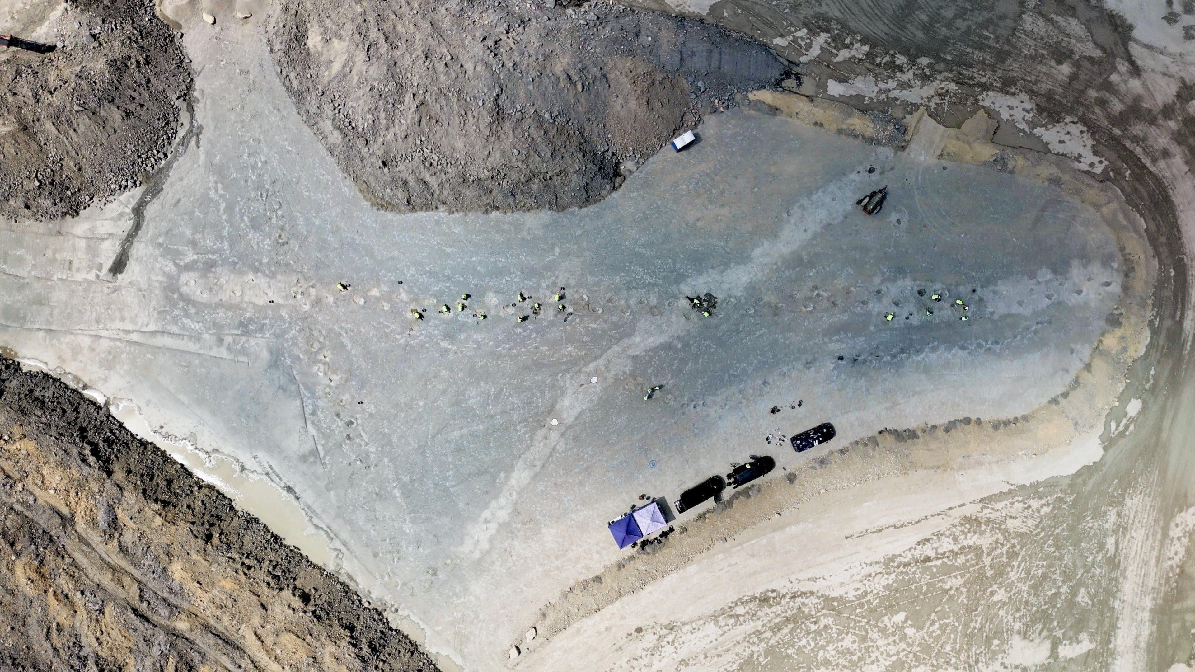 Qn overhead drone shot taken from about 200 metres up shows a large quarry with the two sets of dinosaur prints criss-crossing it. There are also several vehicles, a couple of tents and about 15 workers in yellow hi-visibility clothes.