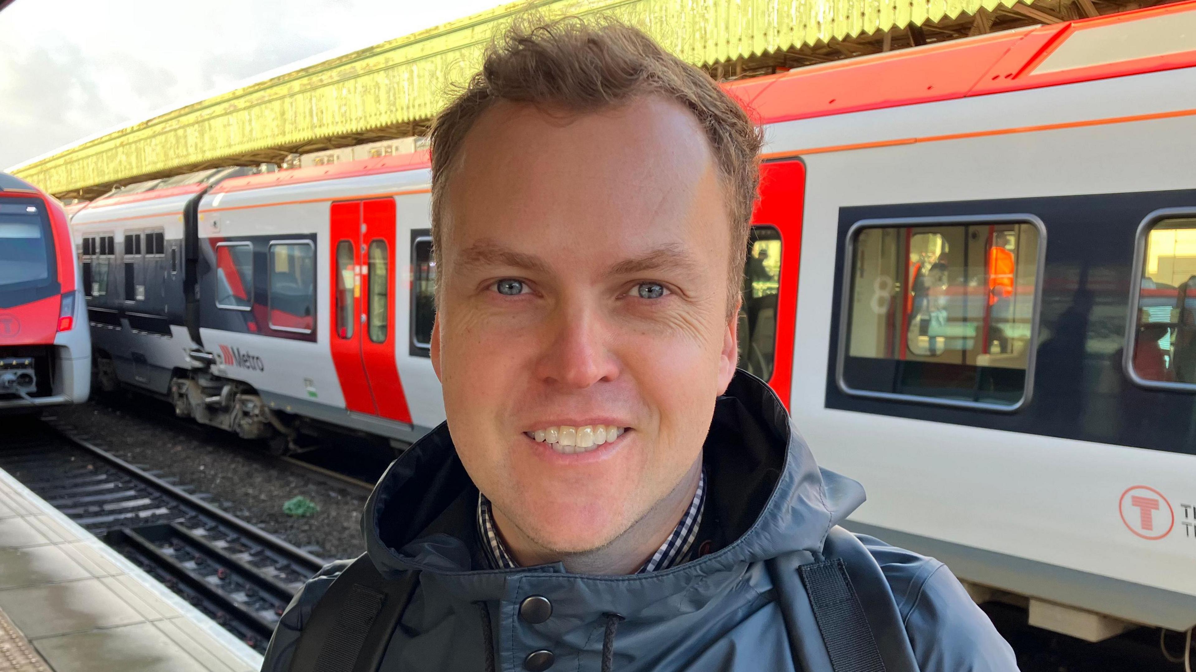 Gethin Jones, swyddog materion cyhoeddus Trafnidiaeth Cymru yn sefyll ar y platform yng ngorsaf Caerdydd Canolog ac yn edrych yn syth i'r camera. Mae e'n gwisgo cot law glas tywyll.