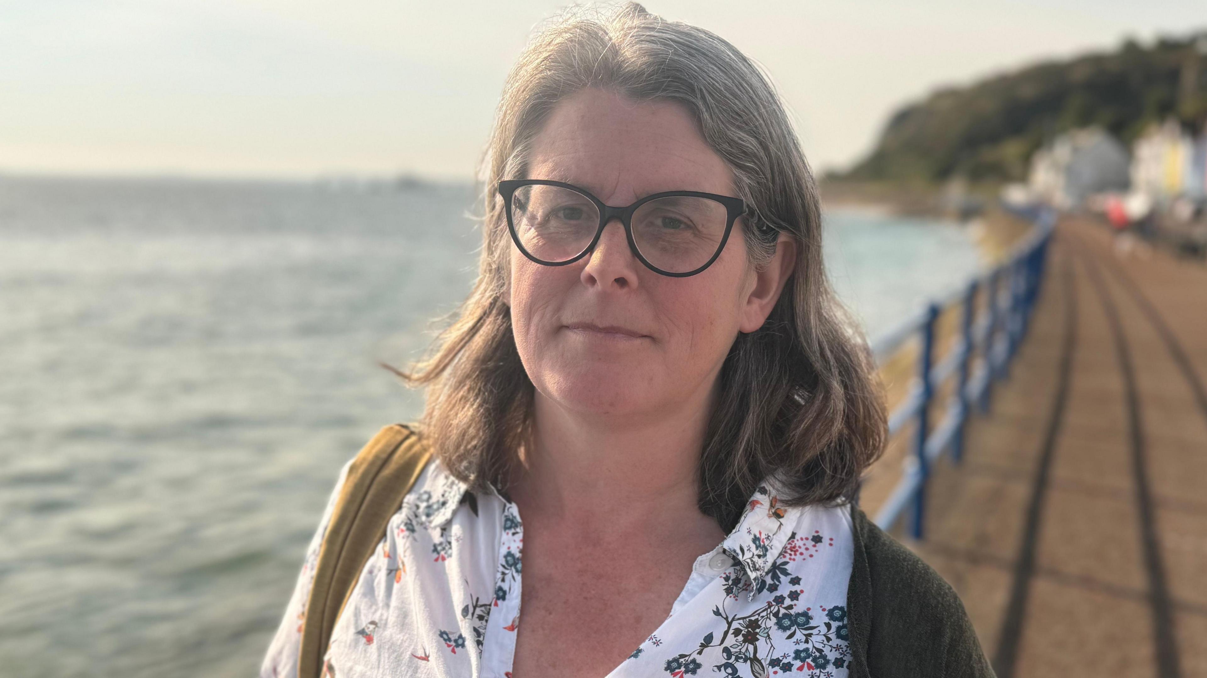 Clodagh Miskelly - a woman with grey, shoulder length hair and black rimmed glasses. She is standing on a promenade beside Belfast Lough and looking at the camera. She is wearing a white patterned shirt