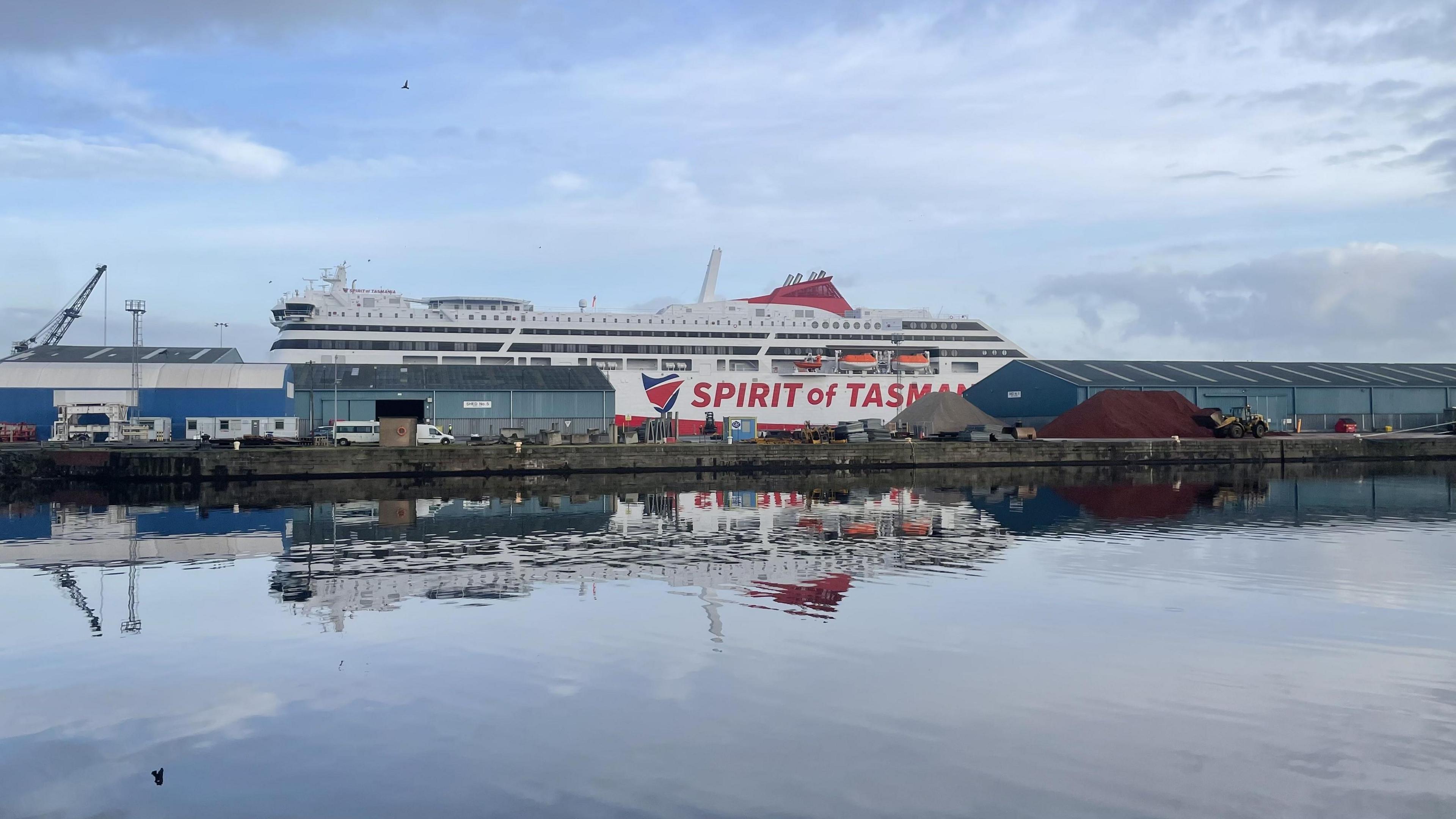 The Spirit of Tasmania ship berthed in Imperial Dock in Leith. The ship is partially obscured by a blue warehouse building. It is mainly white, with a red hull and the words 'Spirit of Tasmania' in red on the side. The picture is taken from a distance, with water in the foreground.