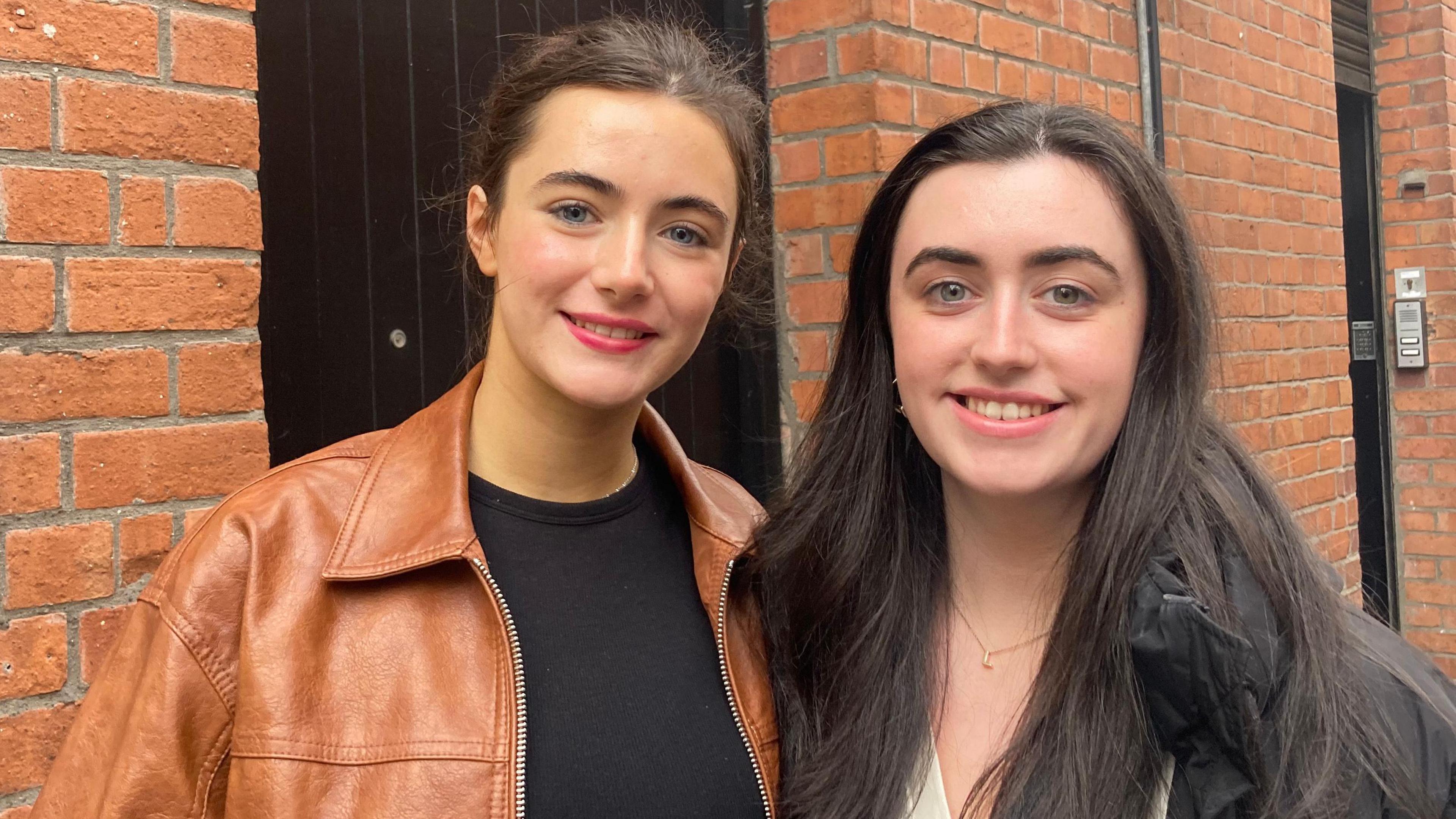Lily Rose and Emily Jane are both wearing jackets. 
They have brown hair and are smiling near a bricked wall. 