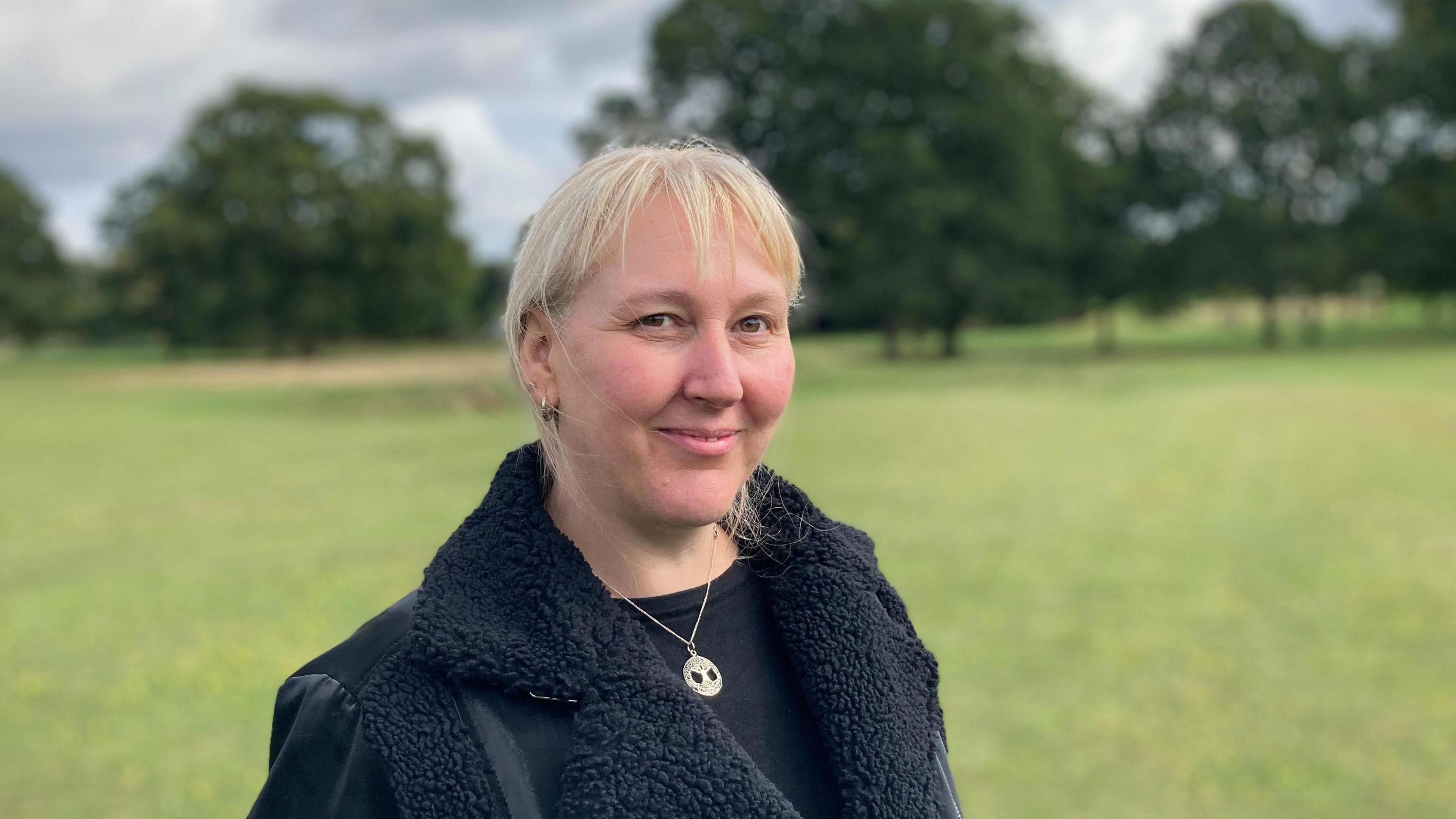 Sammie Wakefield with short straight blond hair, smiling at the camera and wearing a black coat and silver necklace. She is standing in a park with trees in the background.