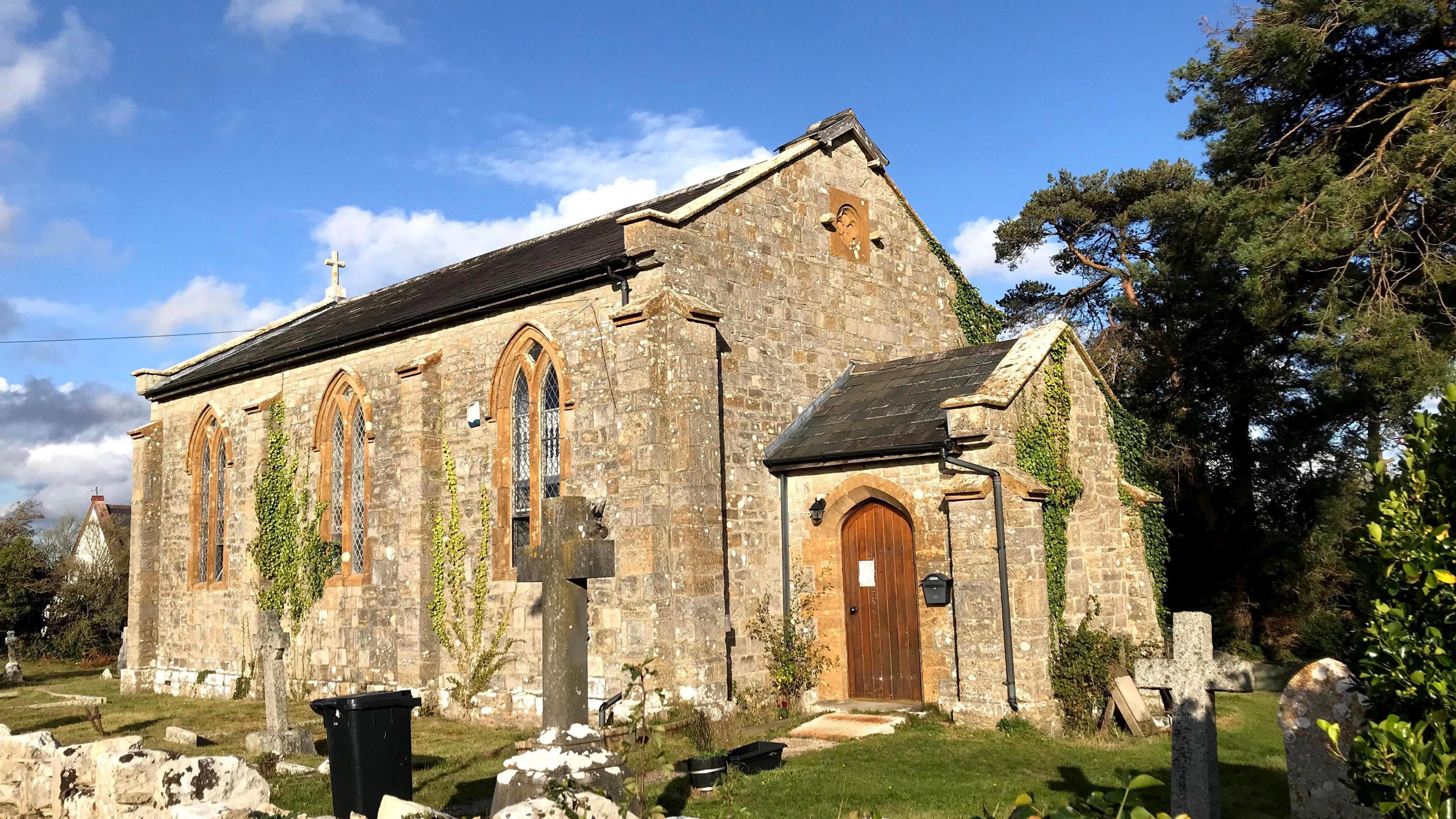 An exterior shot of the chapel used by Burton Chapel Records. It's a sunny day, the sky is blue and there are a few clouds, the chapel is made of pale brown brick and has large arched windows framing the side, with some greenery growing up the building and a wooden panelled door to enter can be seen to the right. In the background framing the chapel are some trees and hedging, and the chapel sits in a small graveyard, with some tombstones in the shape of crosses in vision.