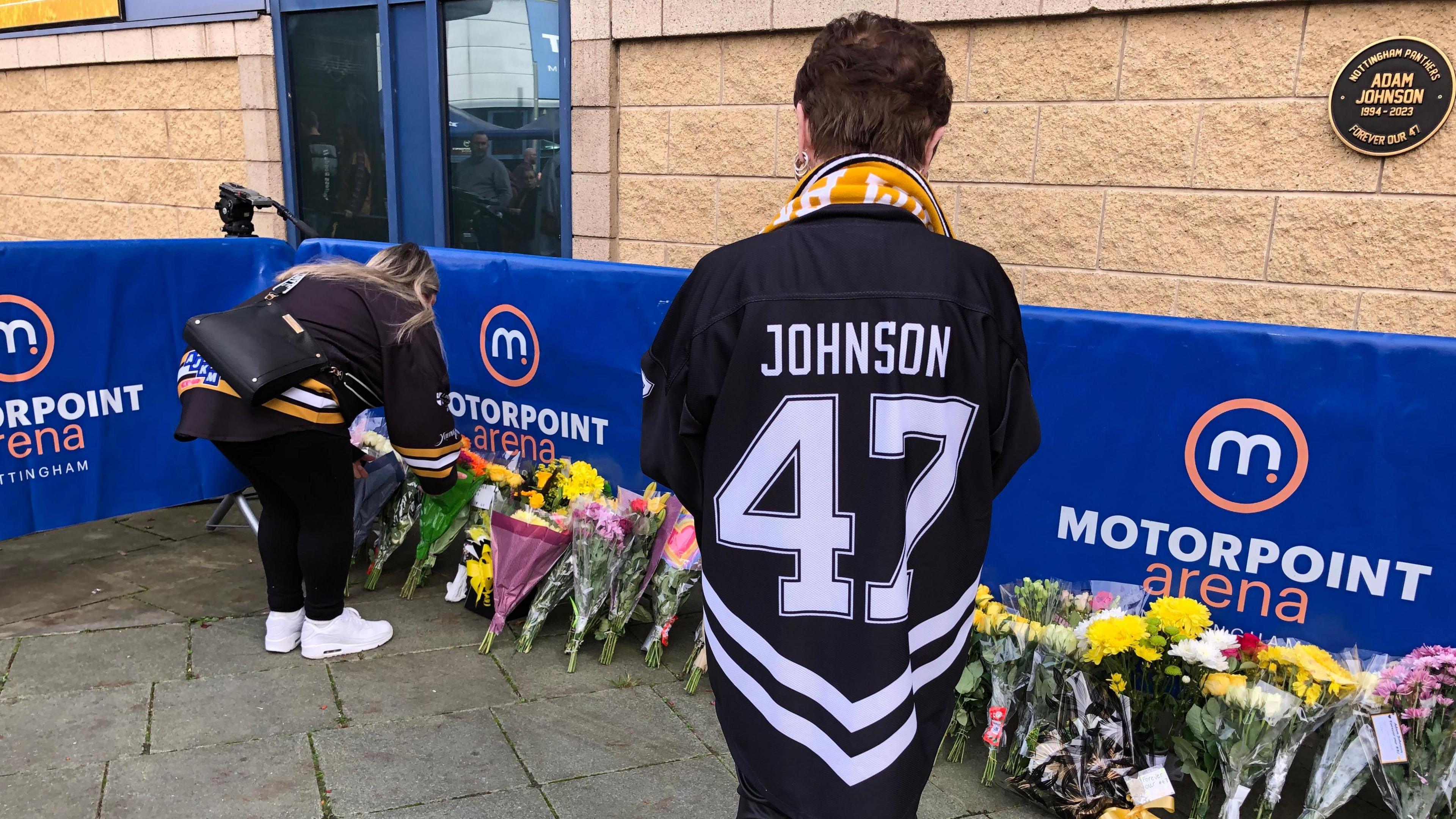 A woman with an ice hockey jersey with "Johnson 47" on the back stood next to flowers in memory of Adam Johnson