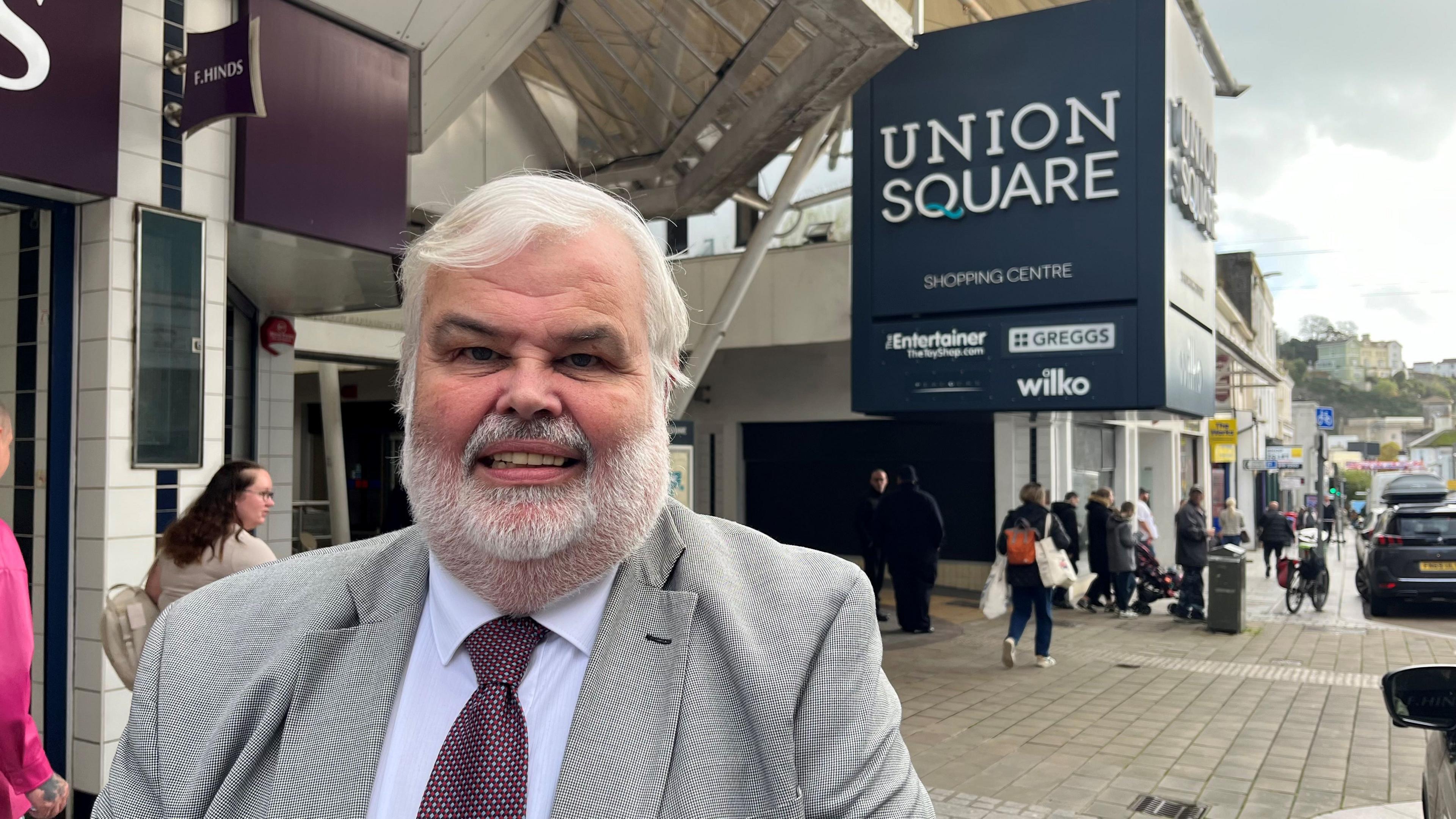 David Thomas, Conservative leader of Torbay Council, wearing a grey jacket and burgundy patterned tie, standing in front of the Union Square sign