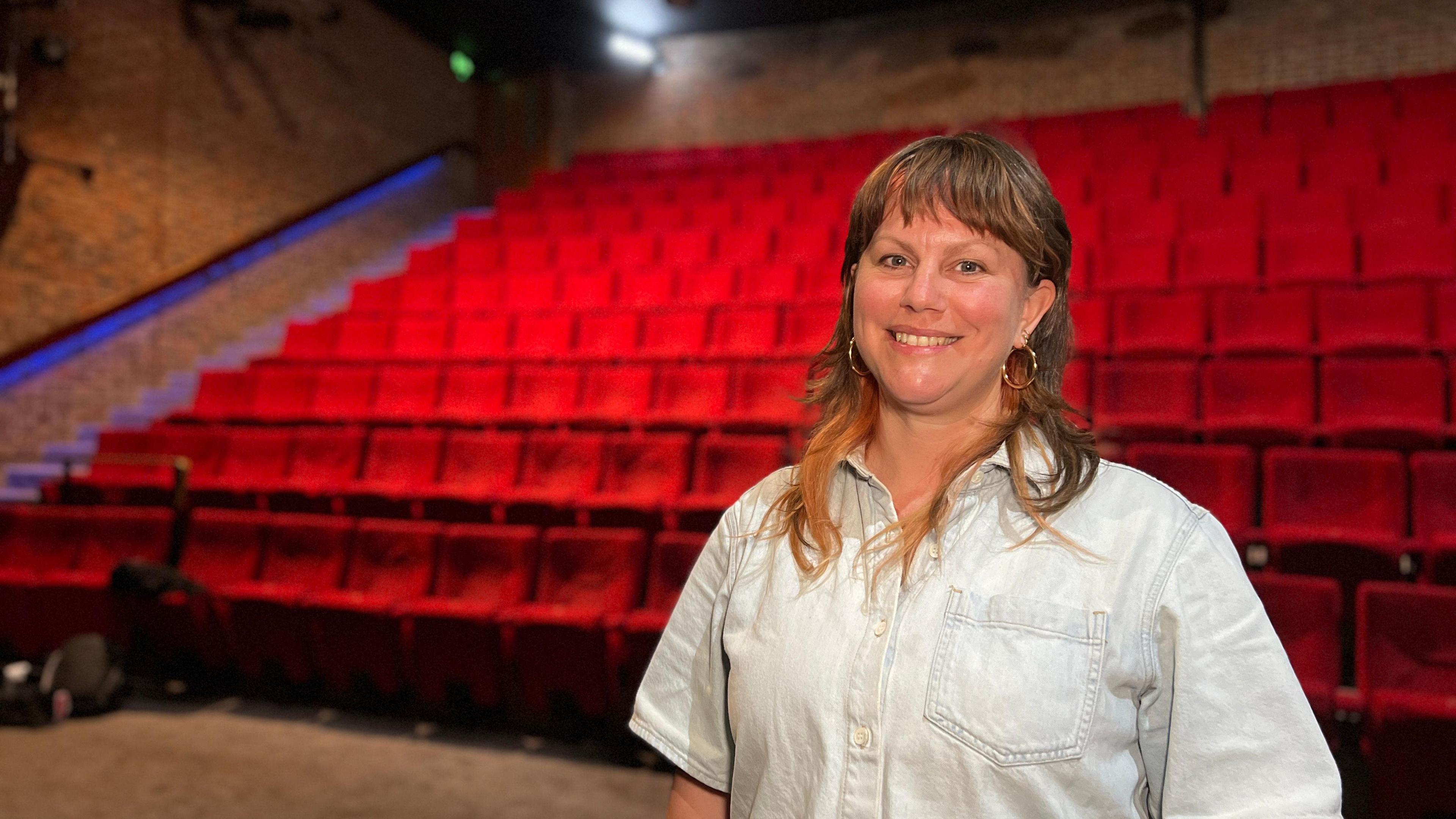 Esther smiles at the camera as she wears a short sleeved, light blue denim shirt. She has short brown hair which comes to her shoulders. Behind her are red theatre seats.