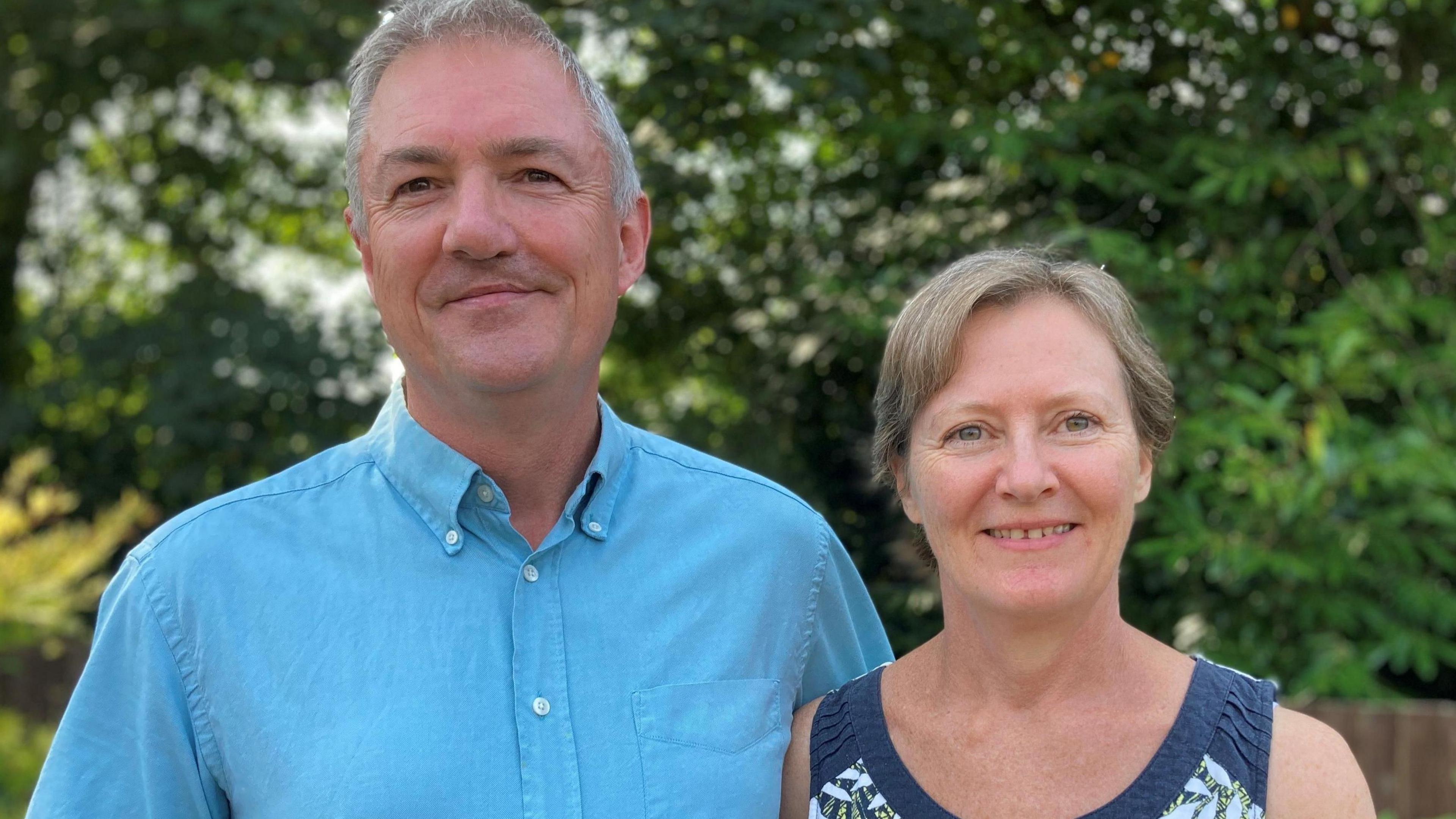 Husband and wife, Stephen and Helen Hull, stand in their garden at their home in Hampshire. Stephen is wearing a light blue shirt and Helen is wearing a strapless blue, white and green floral top.