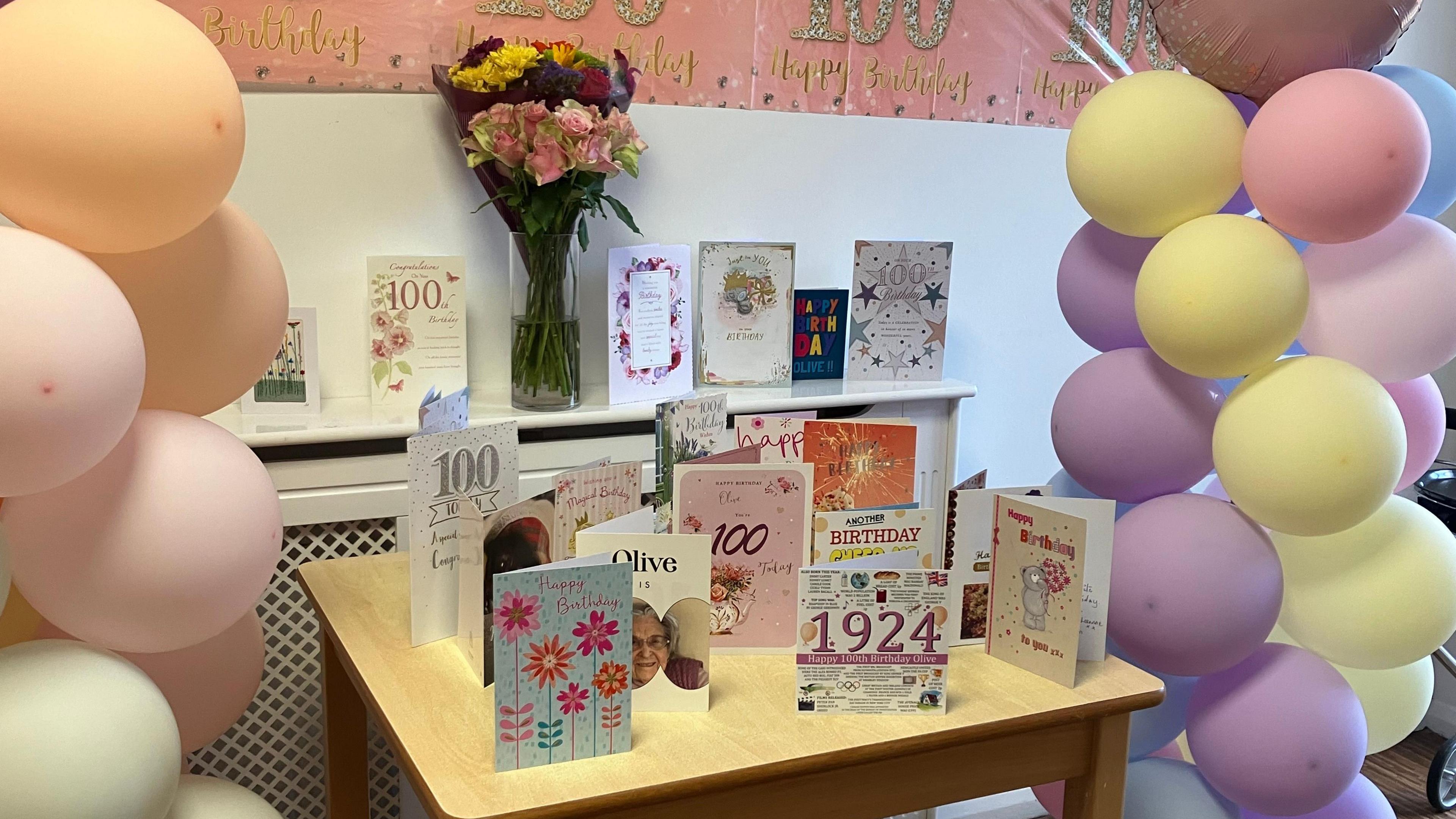 100th birthday cards can be seen on a wooden table with yellow, purple and pink balloons either side. 