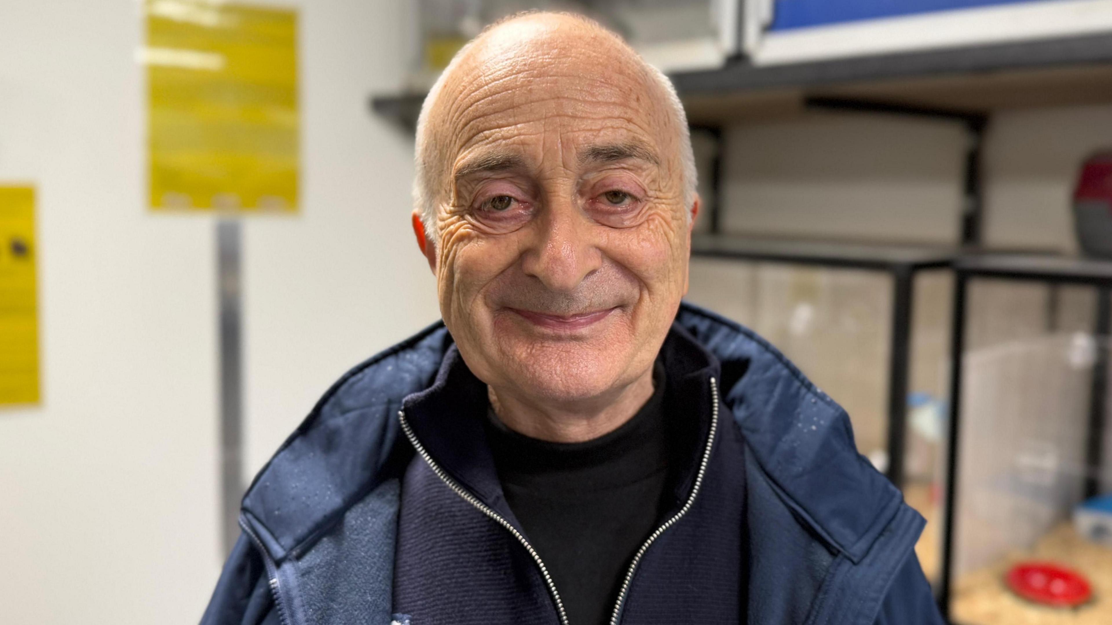 Image of Sir Tony in front of some hamster cages. He is wearing a blue coat and is smiling for the camera.
