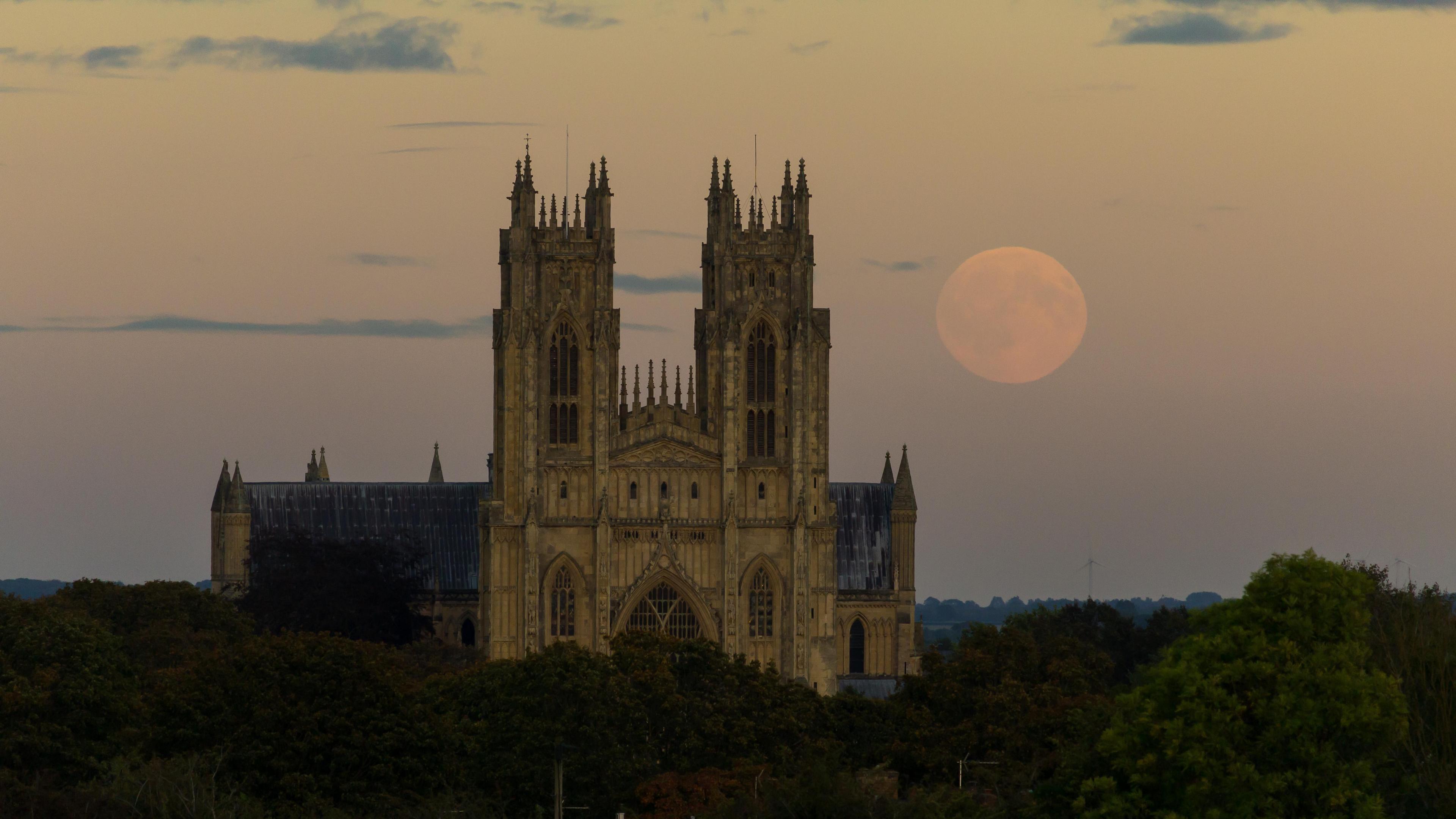 A gothic-style minister with two towers lined with spikes surrounded by green trees, under a sky with a full Moon at sunset. The sky is tinged with pink and yellow hues- and the moon is almost orange in appearance.