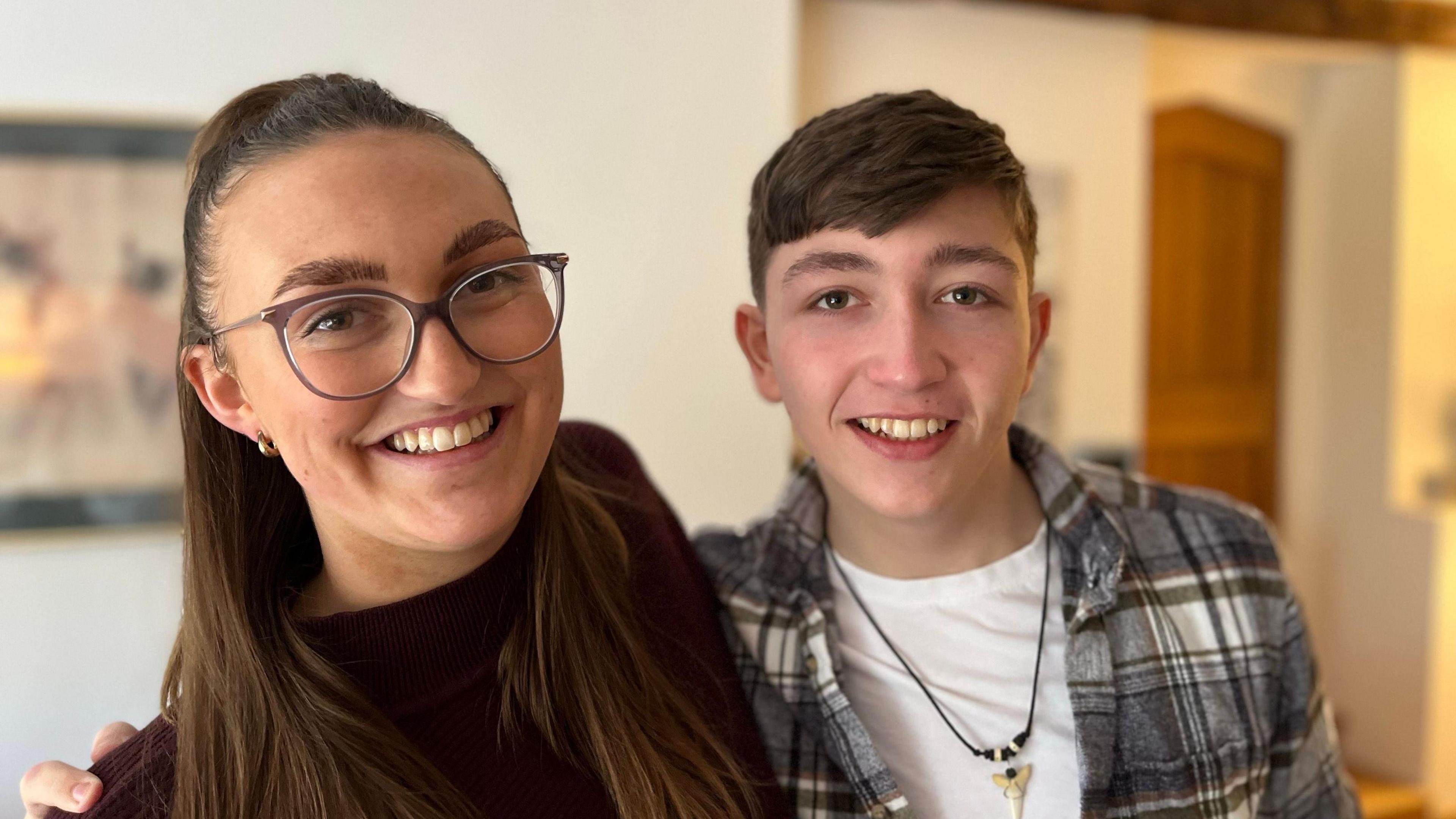 Jayme is wearing glasses and a maroon top with her hair tied back and Ted is wearing an unbuttoned chequered shirt with a white t-shirt with a shark tooth necklace. Both are smiling at the camera standing in a living room 