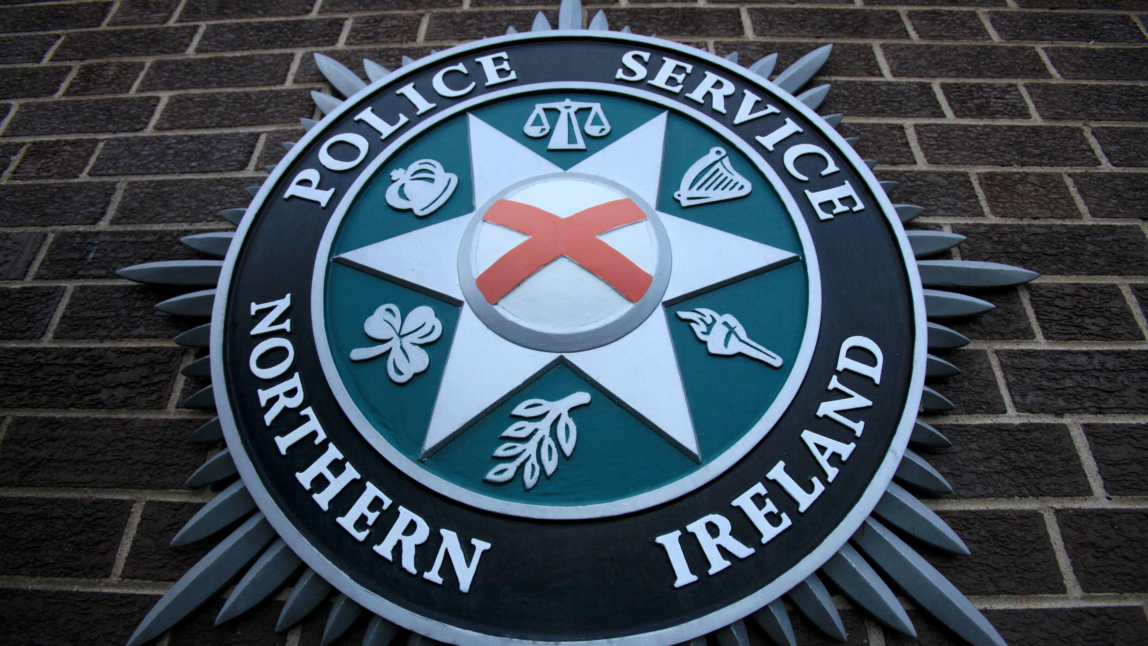 PSNI insignia on a wall. There is the cross of St Patrick and the other symbols are scales of justice, harp and crown from the old RUC badge, the shamrock olive branch and a torch 