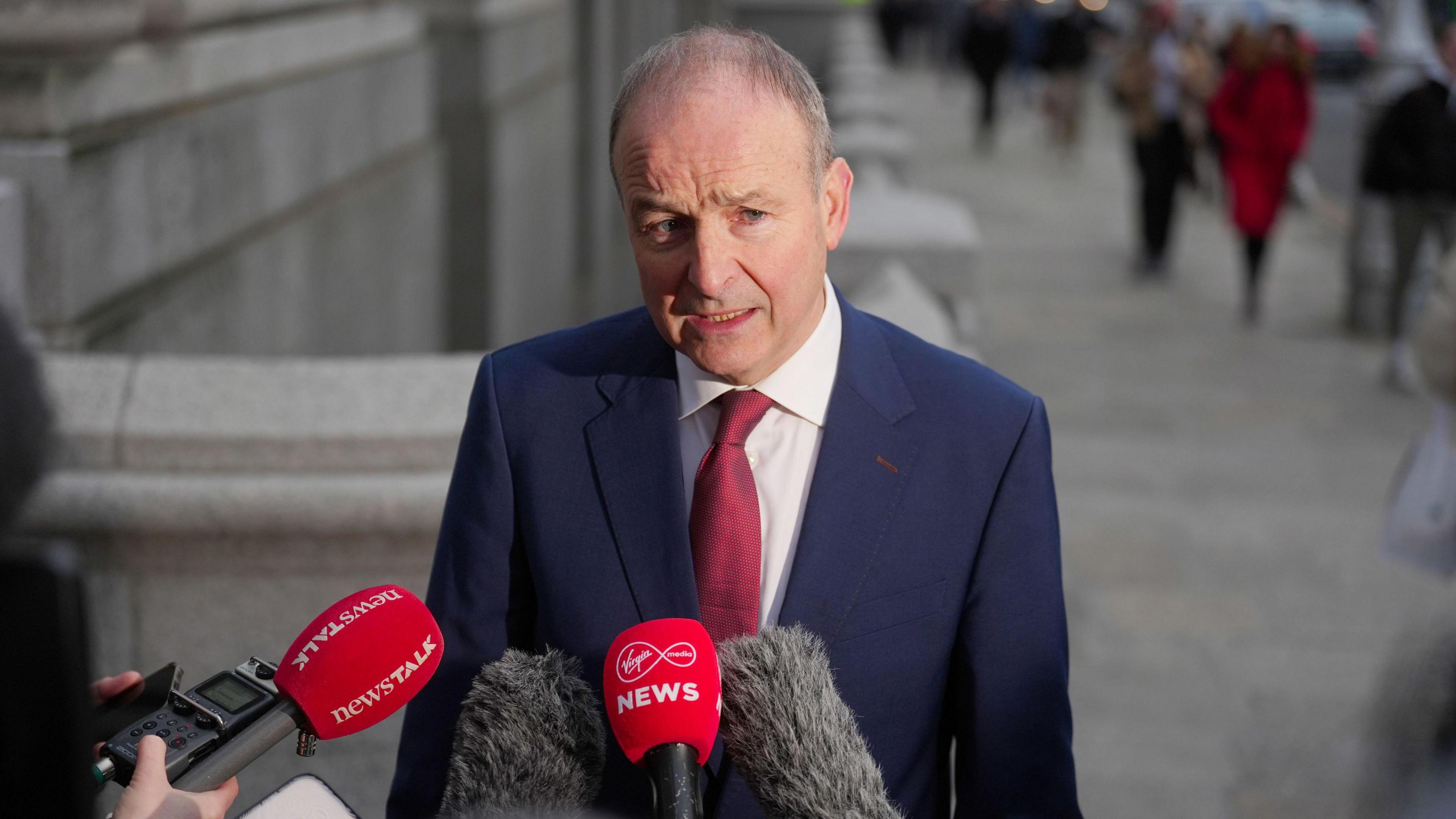 Micheál Martin standing on the street speaking into microphones. One of the microphones has the "NewsTalk" logo on it, another that of Virgin Media News. He is wearing a navy suit and red tie. In the background there are people walking past.