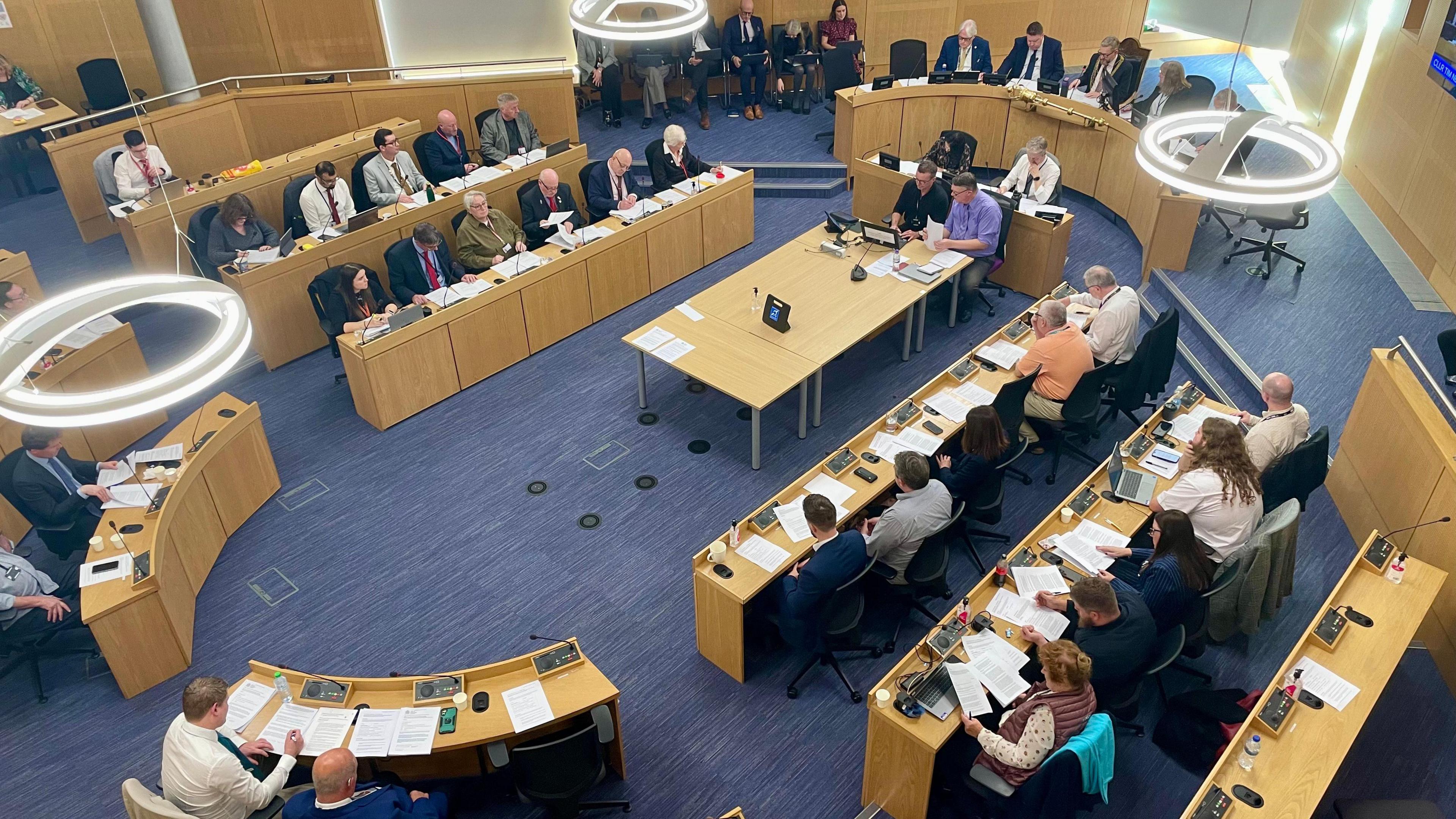 An ariel view of a council room has a number of curved desks with several councillors sitting reading agenda papers