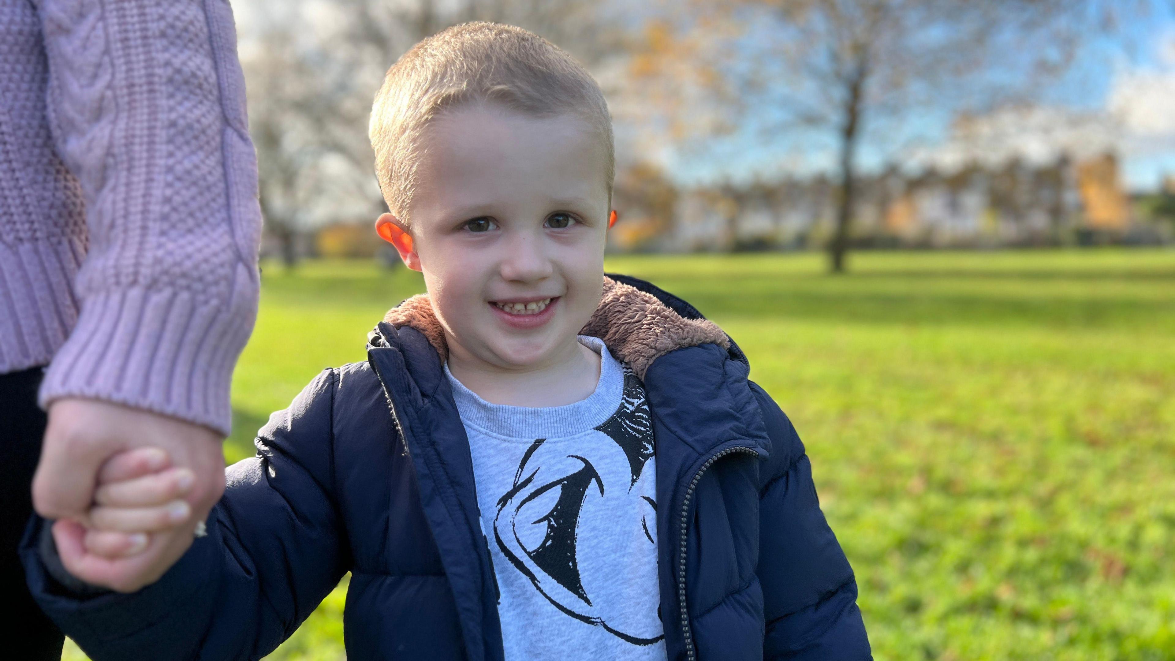 A young boy with blonde hair and wearing a grey Mickey Mouse jumper and a blue puffer jacket. He is smiling at the camera and holding the hand of a woman who is out of shot. There is a large swathe of grass and some trees in soft focus behind him.