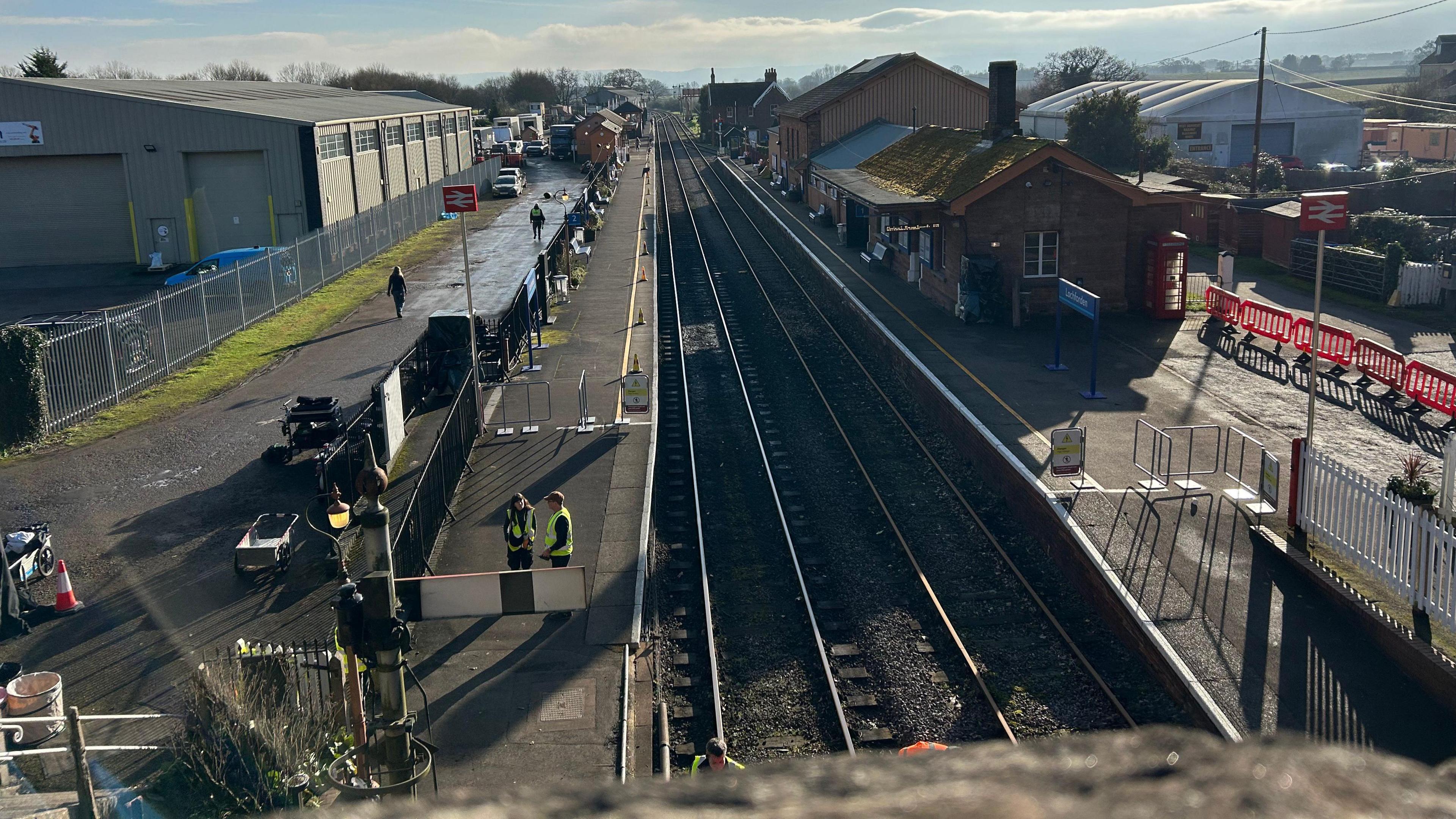 The view of a railway station from a bridge above it. The two stretches of track run out into the distance, while on the left is an uncovered platform. Two people in hi-viz vests can be seen manning a cordon on the platform. On the right is a station building, with barriers beyond that.