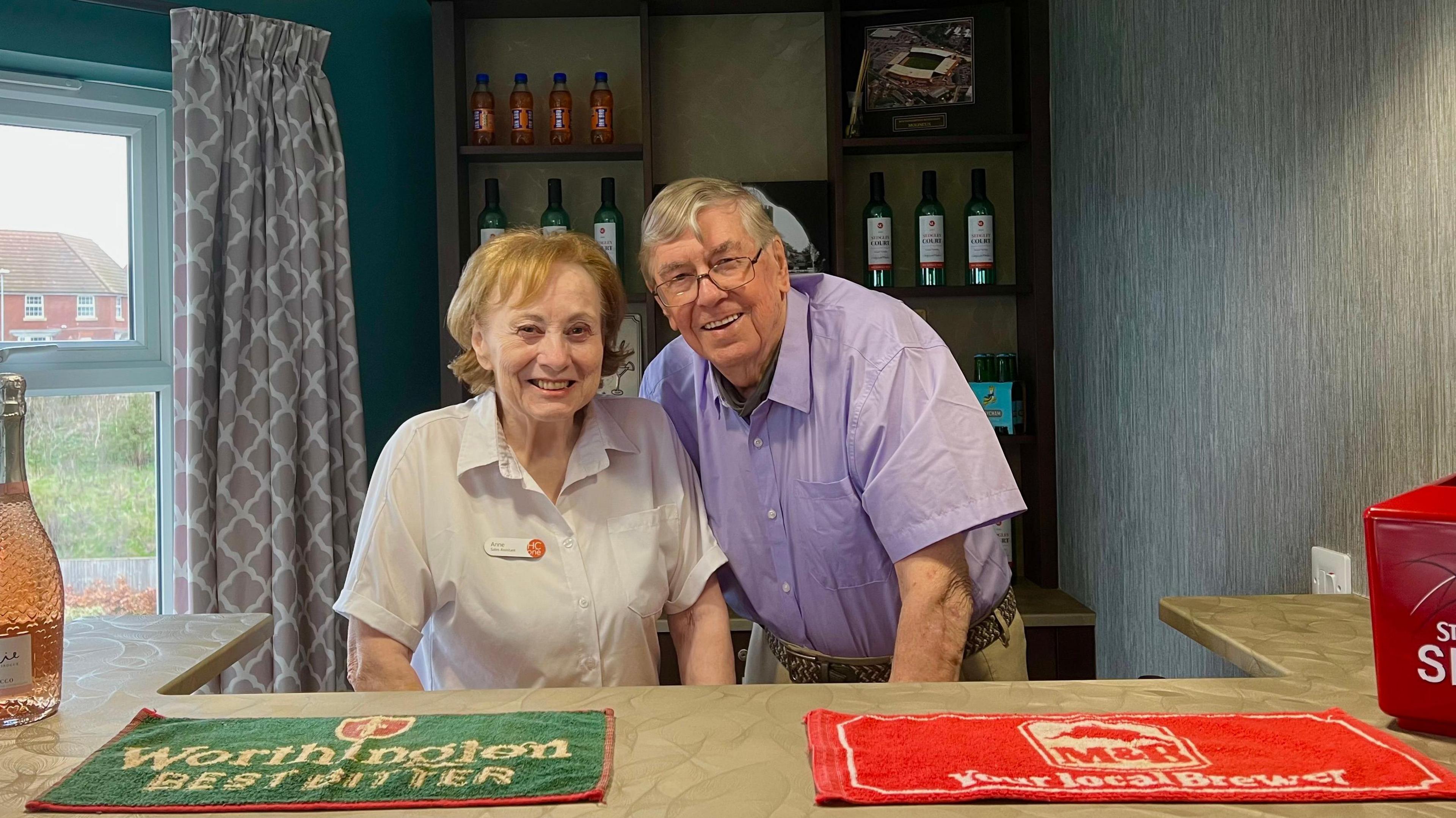 Two people standing behind a bar smiling into the camera. The woman has a white shirt on and the man has a purple shirt on. The man has glasses and grey hair and the woman has ginger/silver hair. 