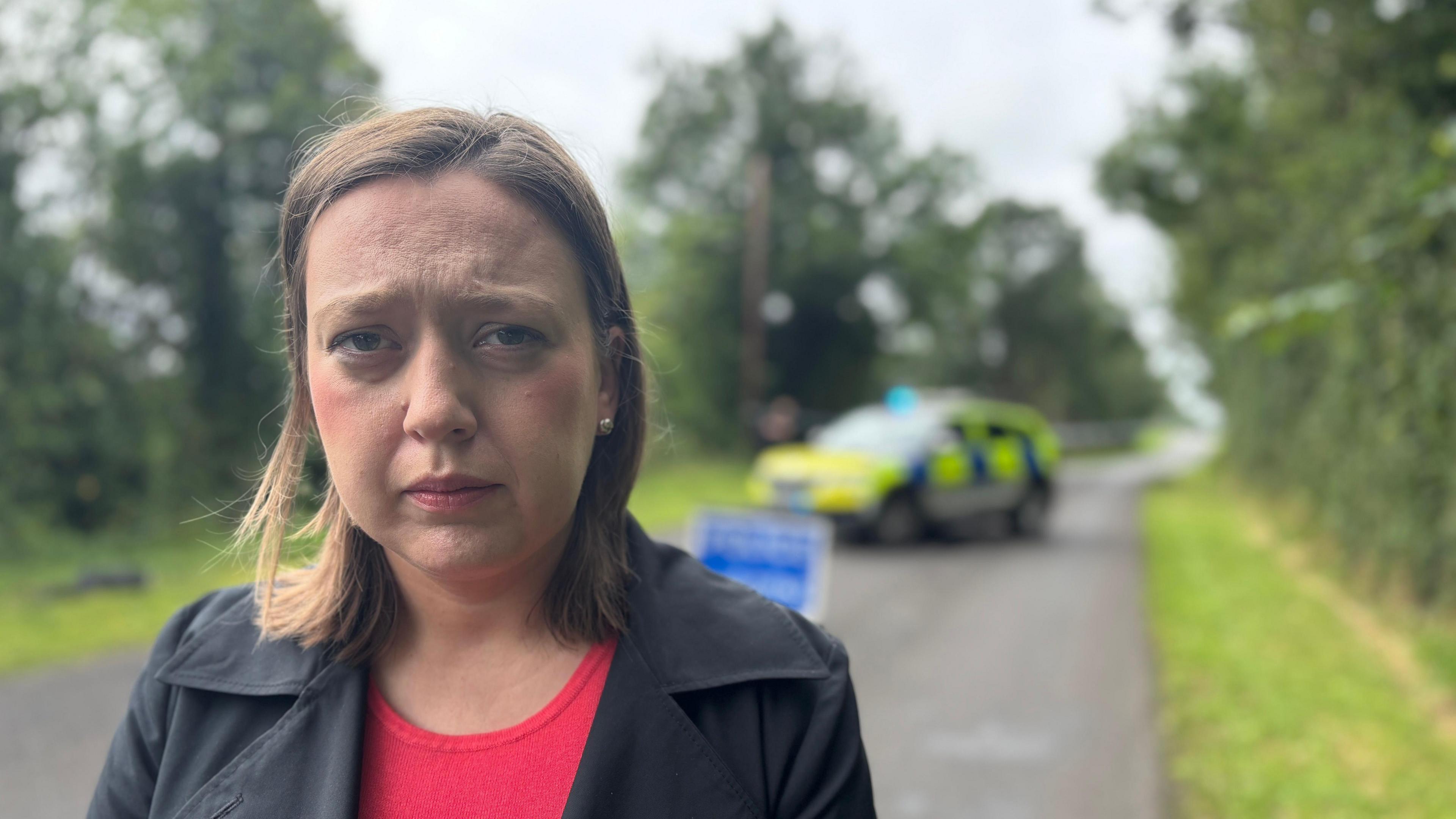 Deborah Erskine stands on the road in front of the police cordon. The police car in the background is out of focus. She is wearing a red top with a black jacket over the top. Her light-brown hair is shoulder length and she has a somber look on her face.