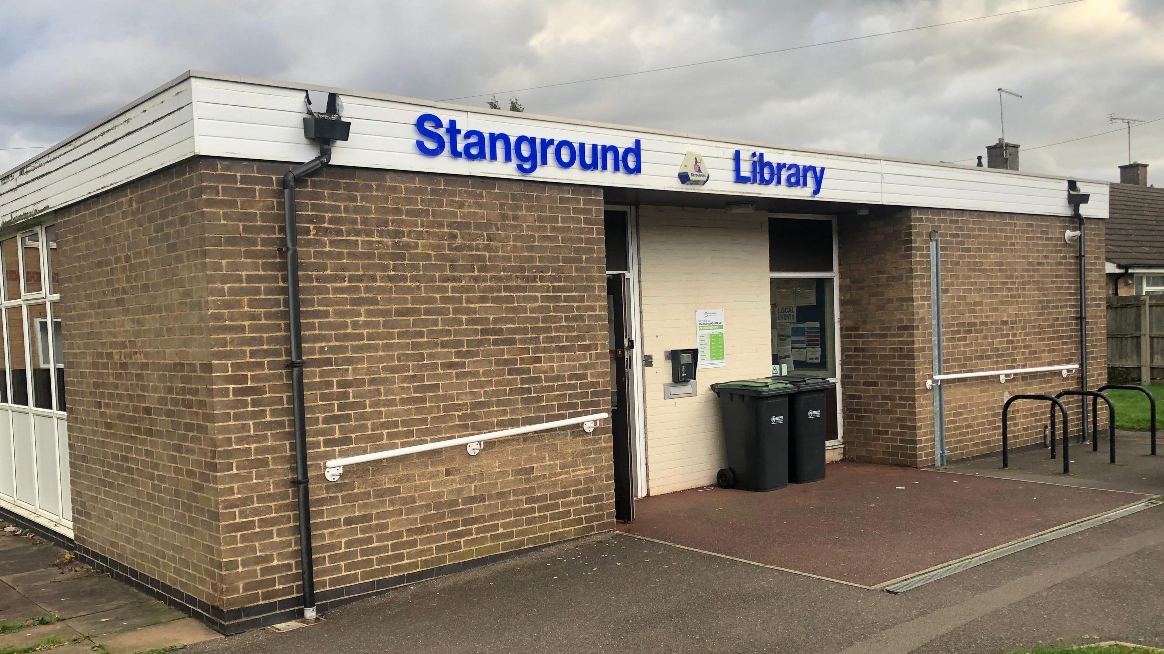 The exterior of a library in Peterborough. It is a single-storey brick built building with a white flat roof. The entrance is in the middle of the wall. Above the entrance it says Stanground library in bright blue capital letters.