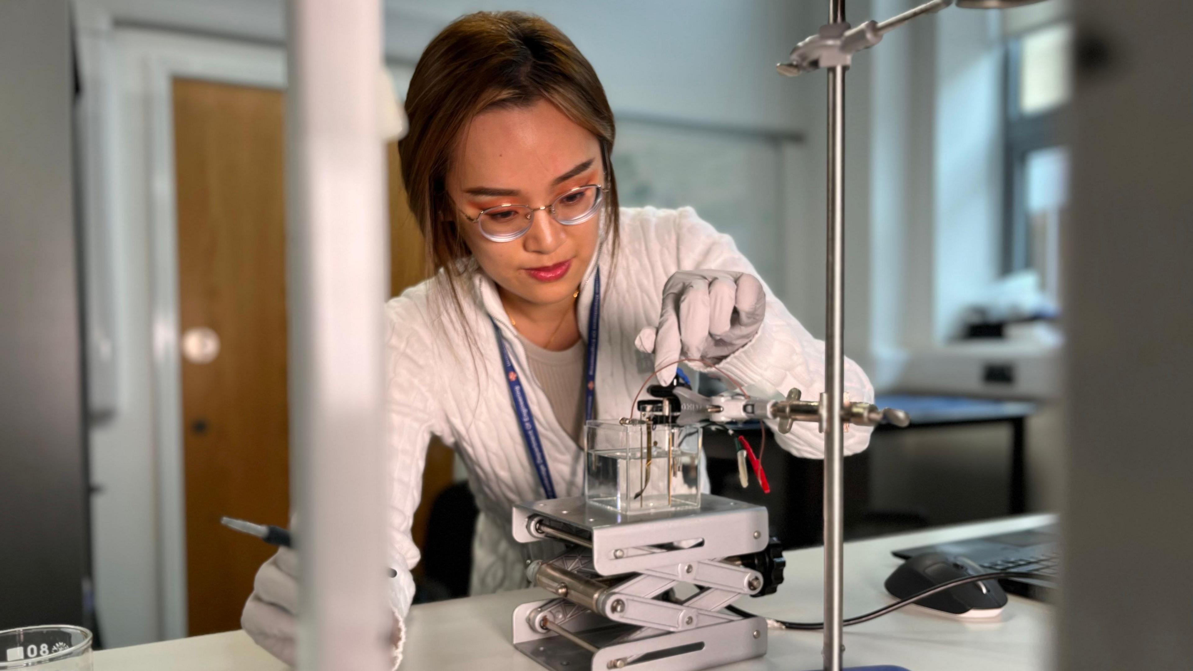 Dr Dong applies an electric current to an implant protoype. She is wearing white gloves while she operates the device. She has long brown hair, circular framed glasses and a white cardigan. 